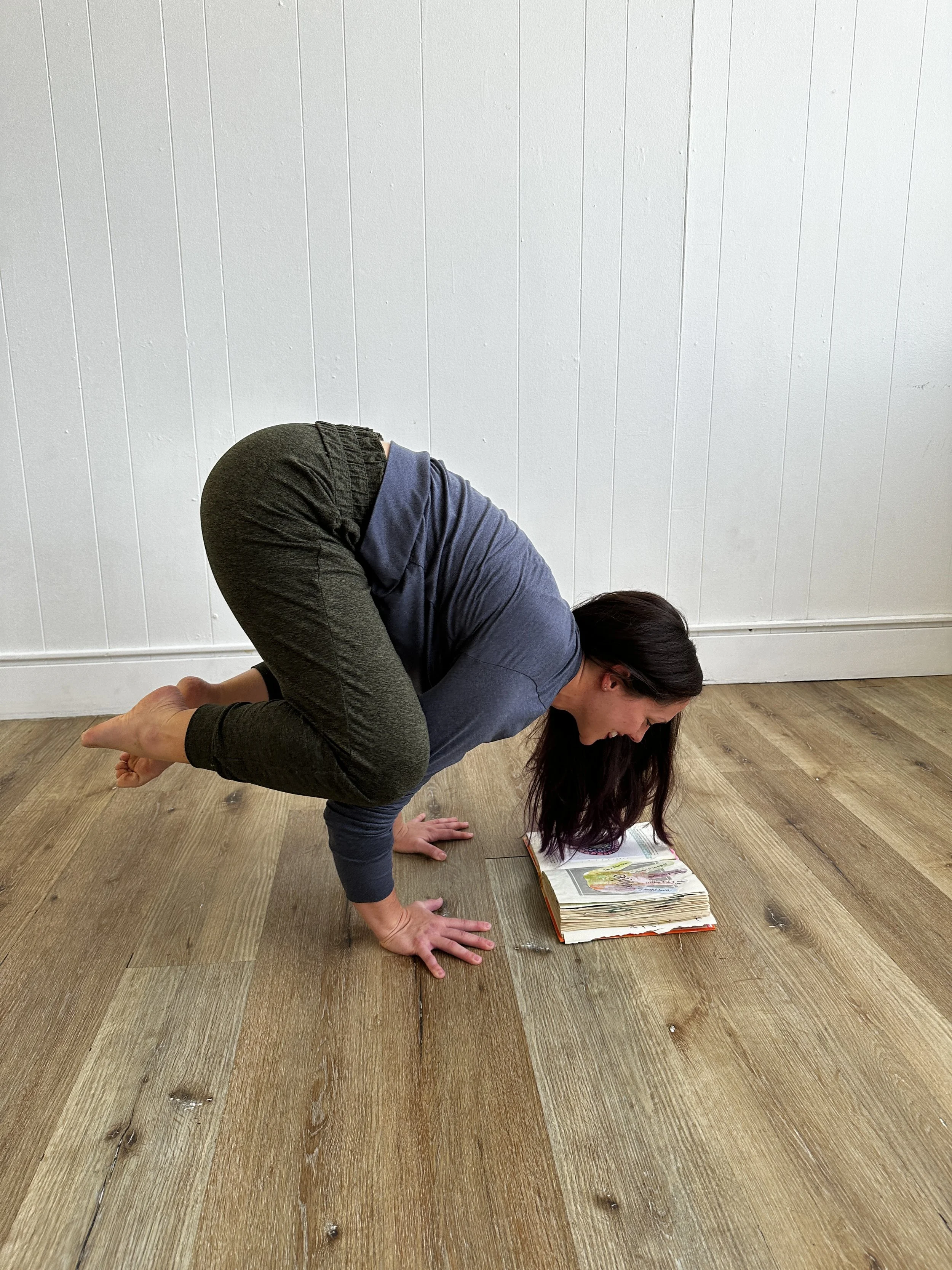Person practicing yoga crow pose on a wooden floor with a book on the ground.