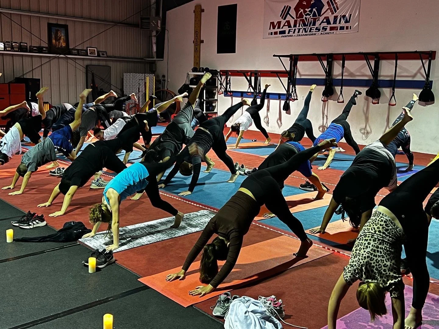 A group of people practicing yoga in a fitness studio, performing the downward dog pose with one leg lifted. The room has exercise mats, candles, and fitness equipment. A banner on the wall says "Maintain Fitness."