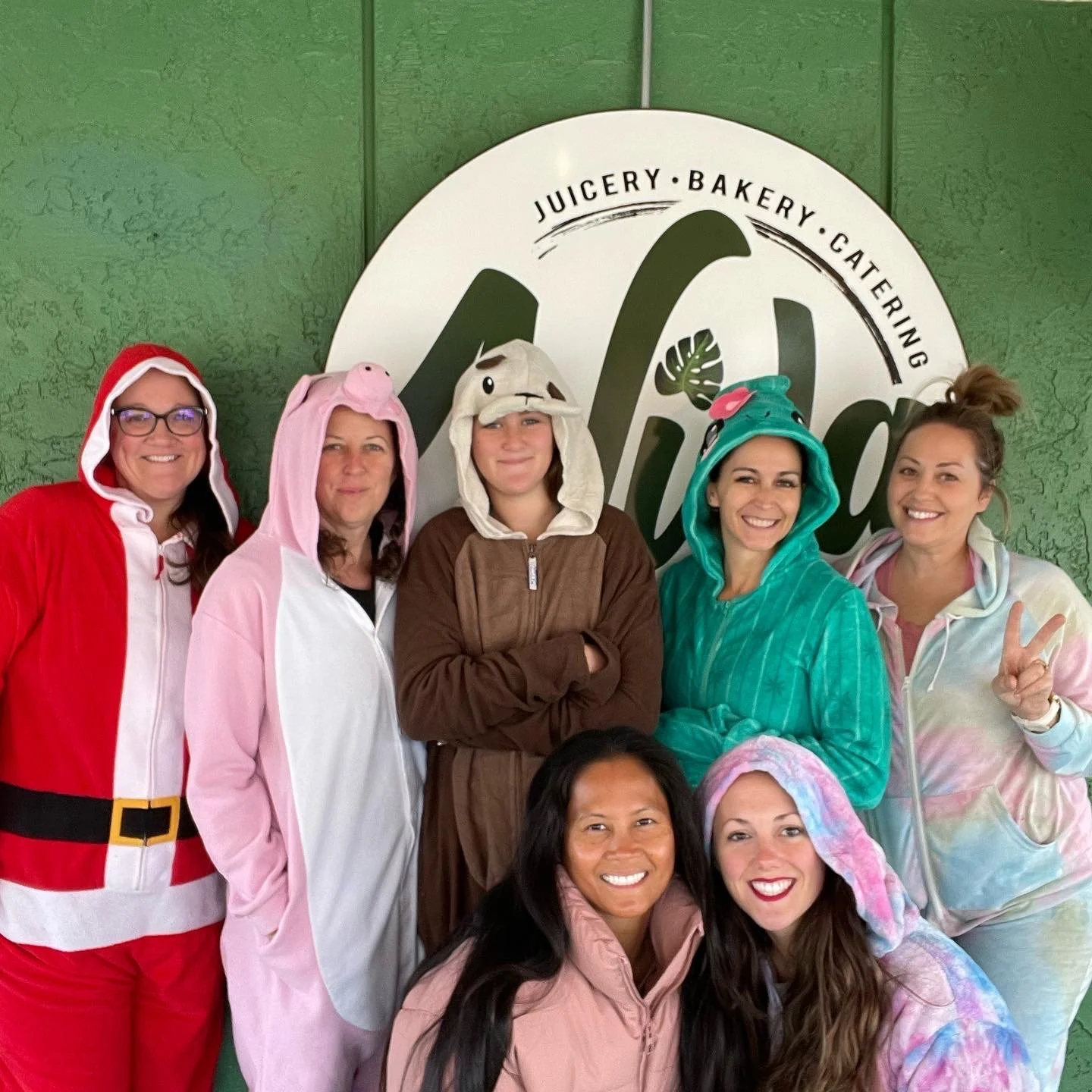 Group of people posing in front of a 'Juicery Bakery Catering' sign. They are wearing colorful animal-themed onesies and costumes, including a Santa outfit, a unicorn, and various animal designs. Everyone is smiling.