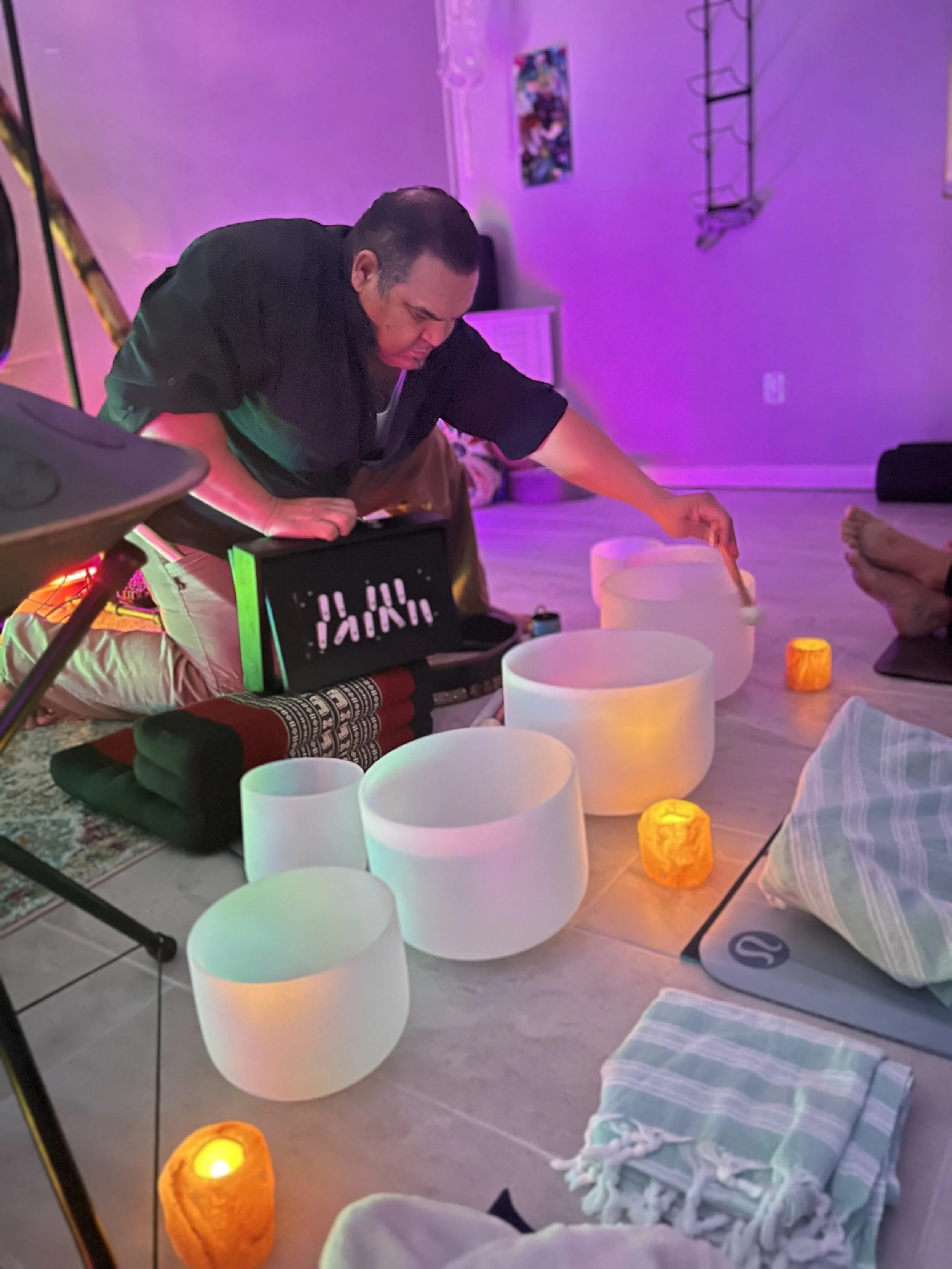 Person playing crystal singing bowls surrounded by candles and blankets in a dimly lit room.