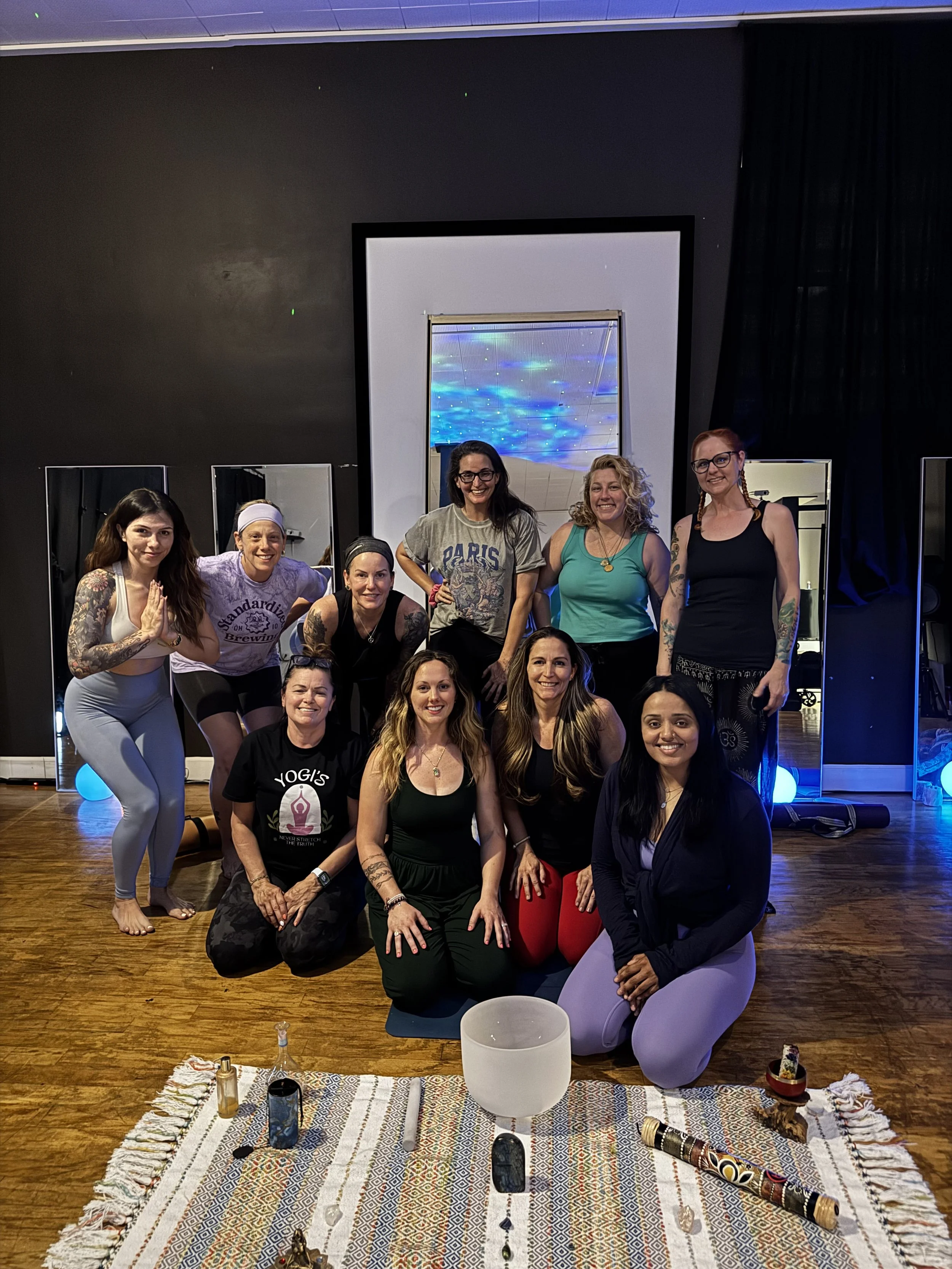 Group of ten women in yoga or meditation studio, some seated on yoga mats and others standing, with spiritual objects and a large singing bowl in the foreground, and a black wall with mirrors and a large screen in the background.