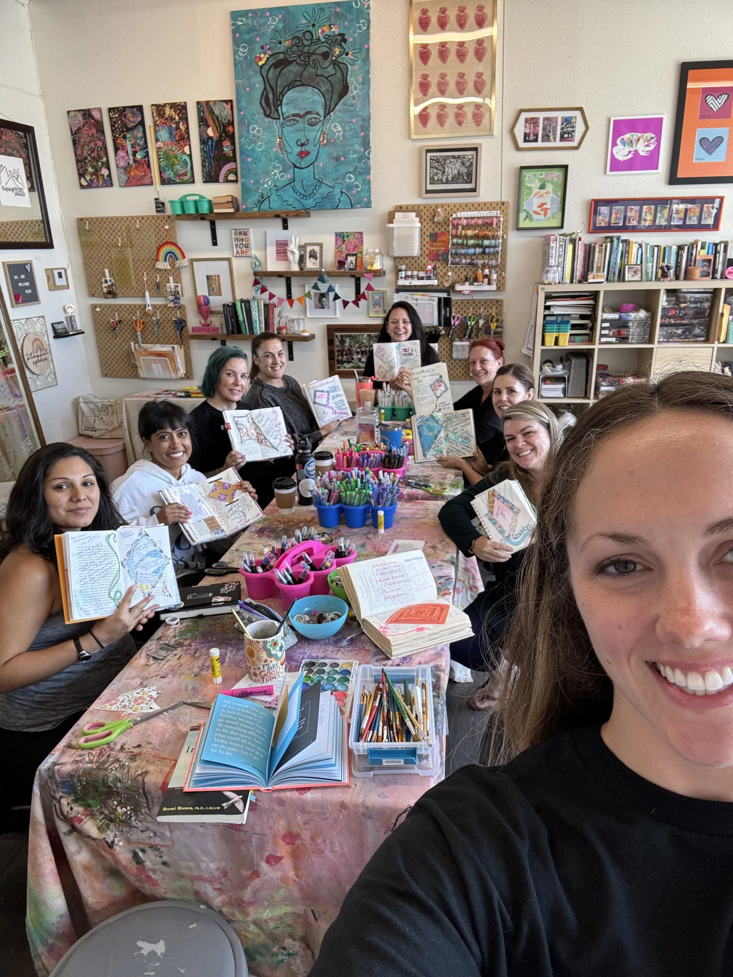 A group of women sitting around a table covered with arts and crafts supplies, smiling and holding open journals or sketchbooks in an art studio decorated with colorful paintings and artwork on the walls.
