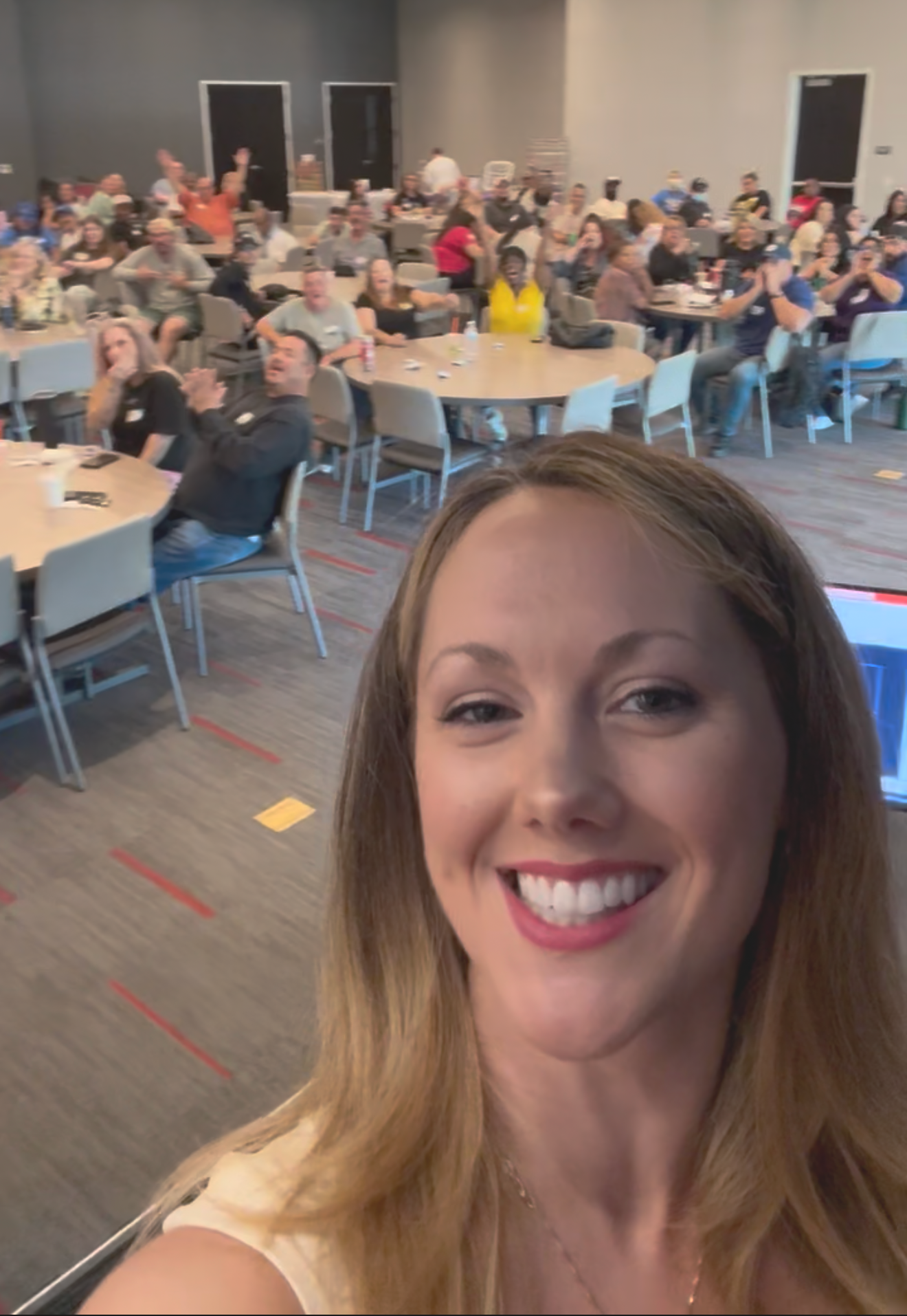 A smiling woman taking a selfie in a large event room with many people sitting at round tables, some raising their hands, others clapping or talking.
