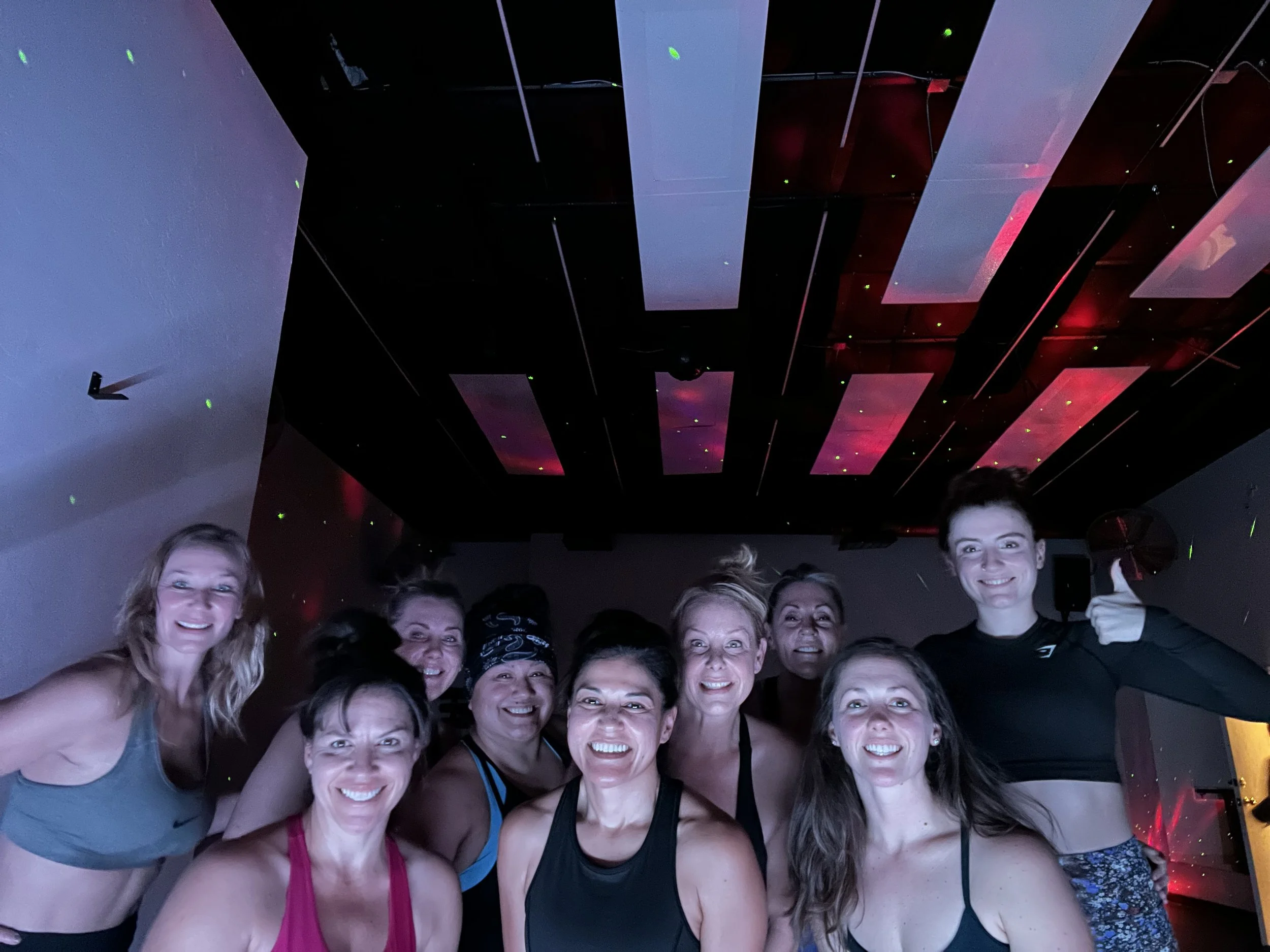 A group of women smiling and posing in a dimly lit room with colored lights on the ceiling.