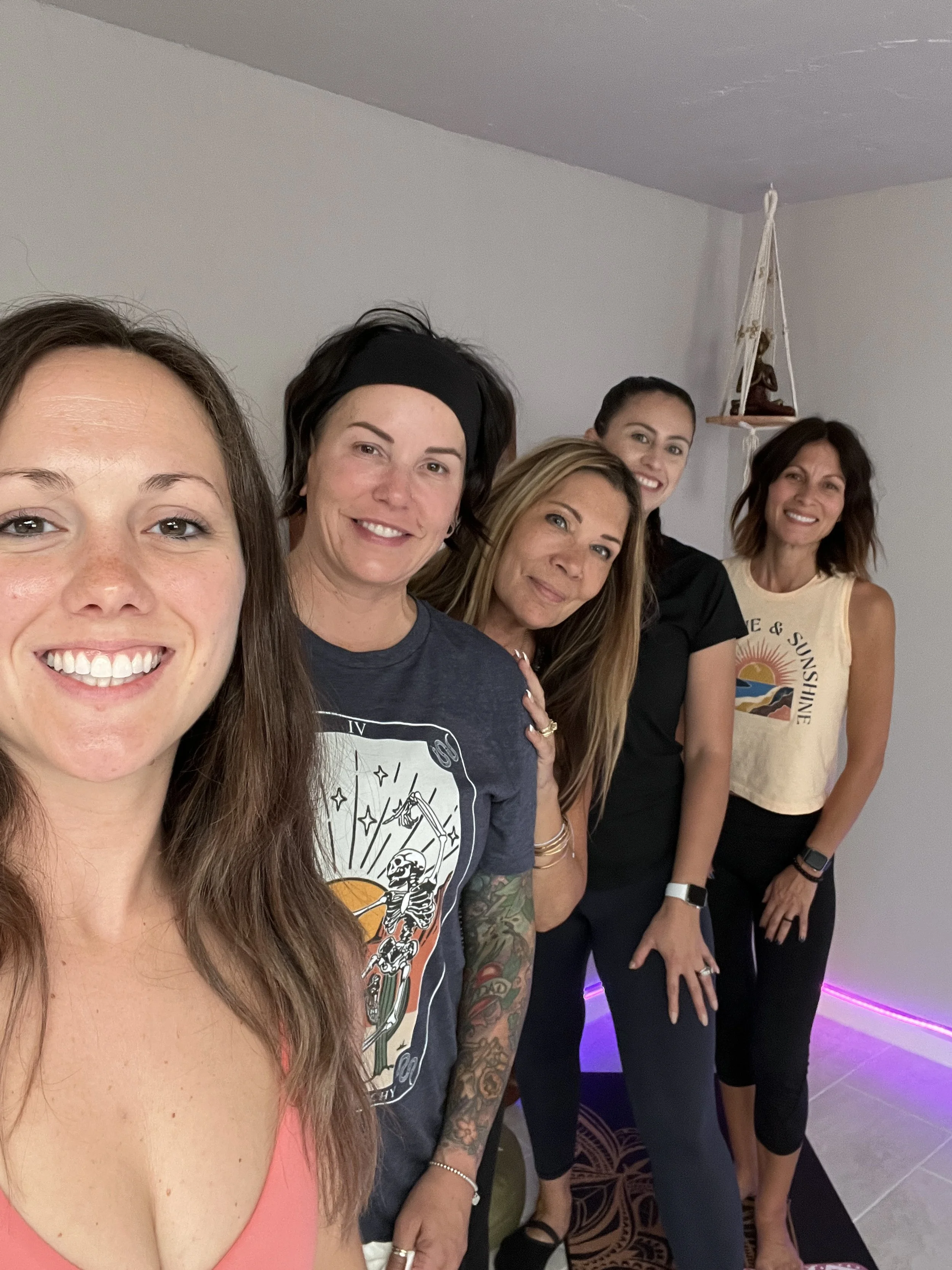 Group of five women smiling indoors, standing together casually with one woman in a pink top at the front. The room has minimal decoration with a hanging shelf in the background.