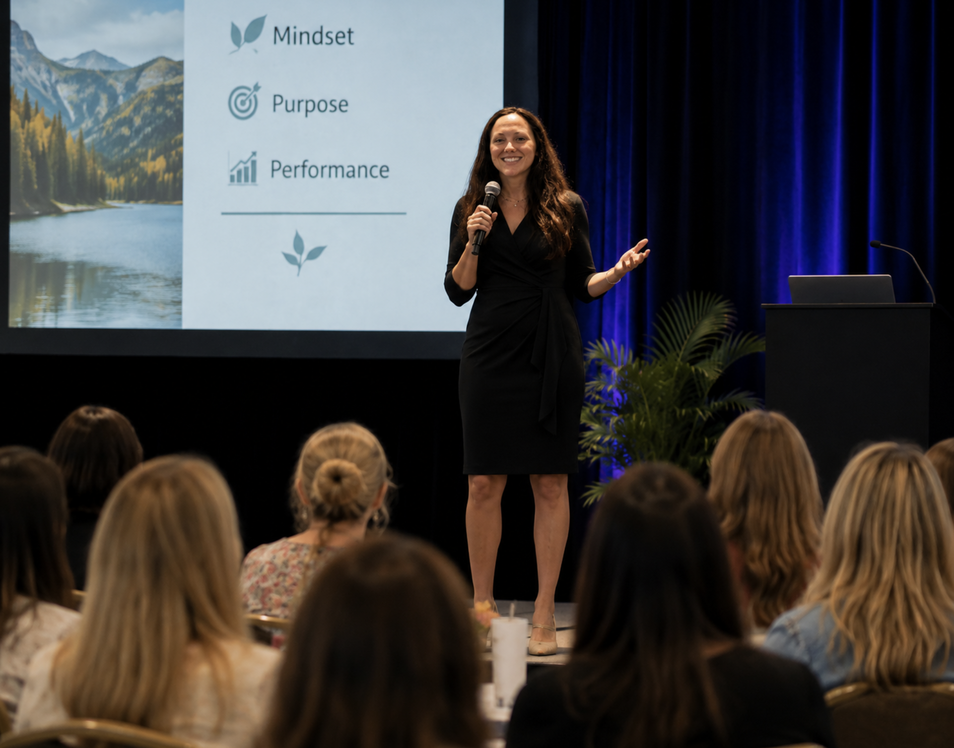 A woman in a black dress is speaking into a microphone on stage in front of a screen that displays words "Mindset," "Purpose," and "Performance" with related icons. An audience of women is seated in front of her.