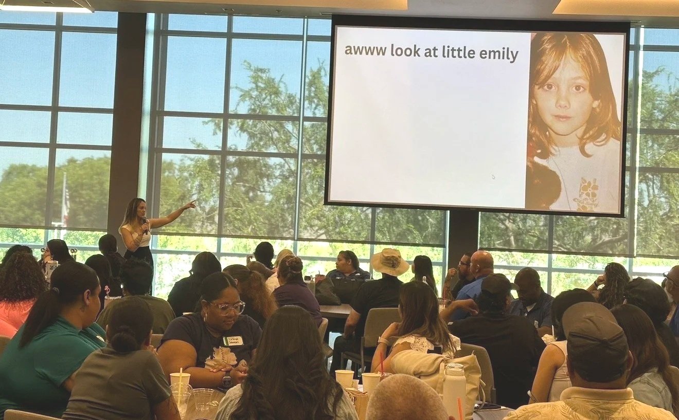 A woman standing on stage pointing at a large screen with a presentation about a young girl named Emily, with a photo of Emily on the right side of the screen. The audience is seated at tables in a bright room with large windows in the background.