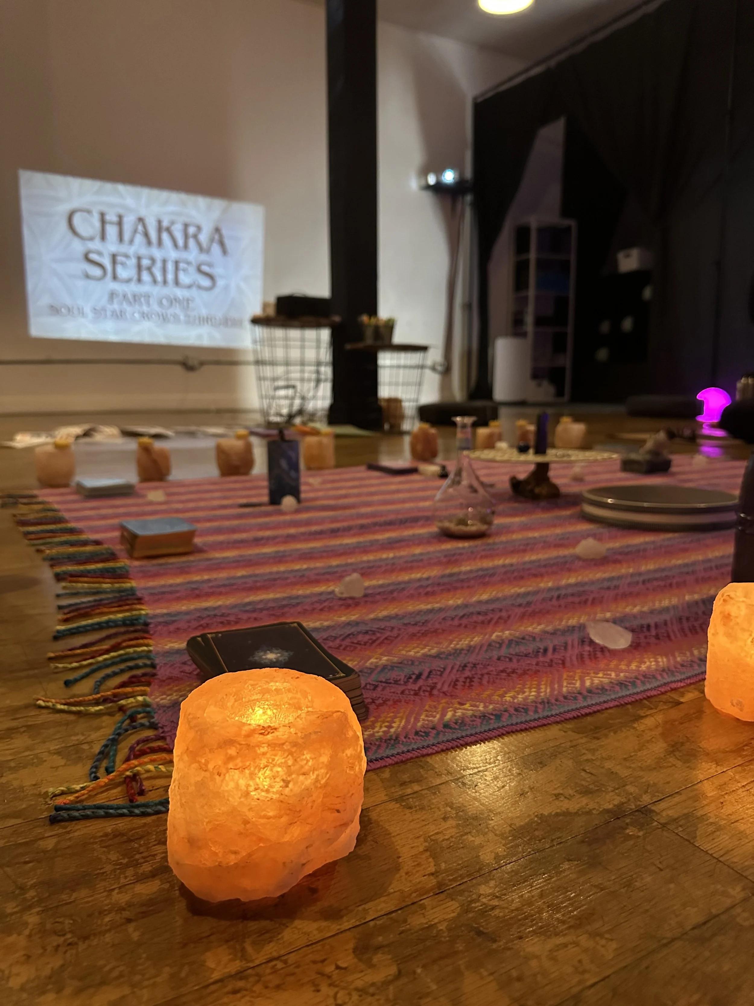 A yoga or meditation session setup with salt lamps, tarot cards, and a decorative cloth on the floor. There is a projection on the wall reading 'Chakra Series Part One, Soul Star & Crown Chakra.'