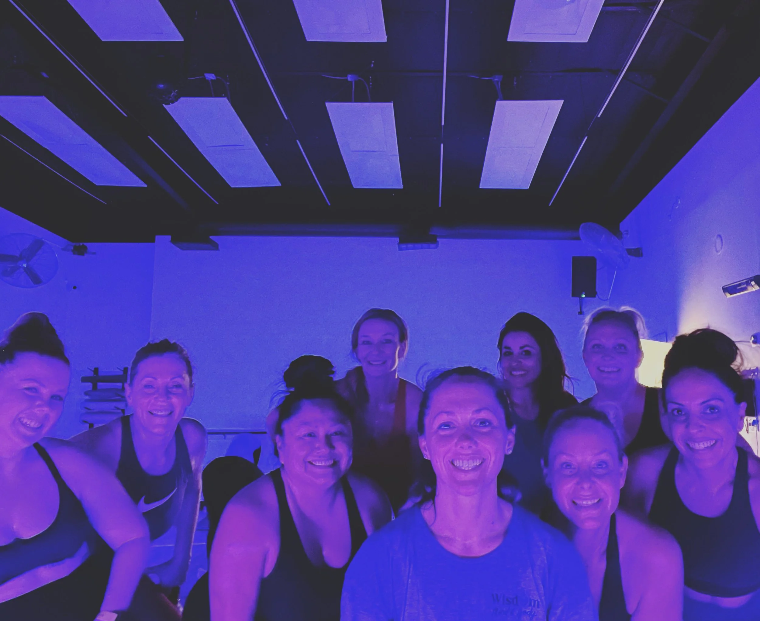 A group of smiling women posing in a dimly lit room with blue lighting, wearing workout attire.