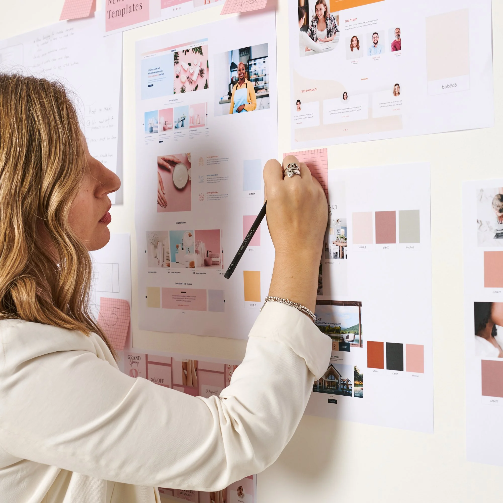 Person reviewing design mockups on a wall, using a pencil to make notes. The wall displays various layout designs, color palettes, and images clipped to it, presumably in an office or studio setting.