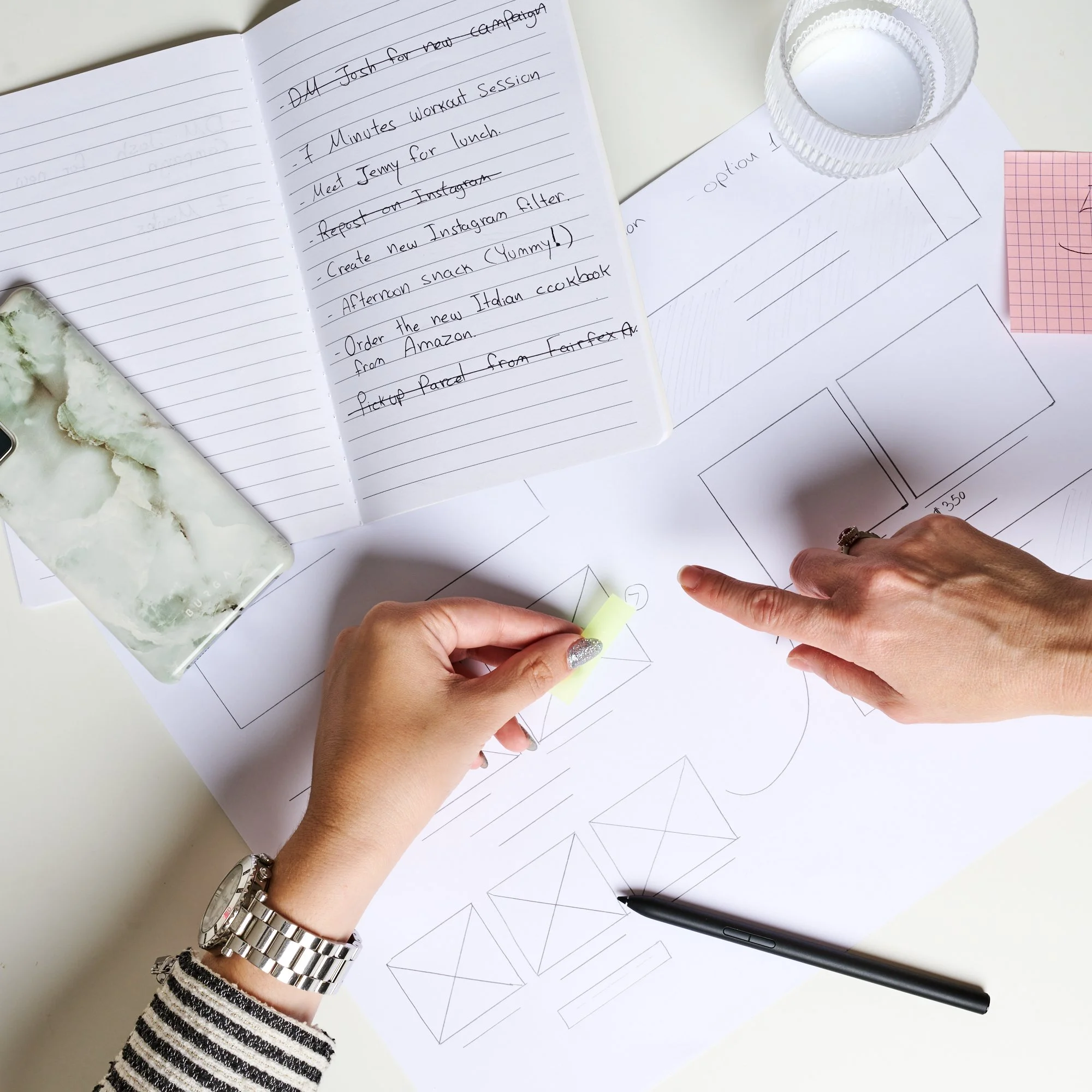 Hands working on a design layout on paper with a notebook listing tasks, a smartphone, glass of water, and pen on the desk.