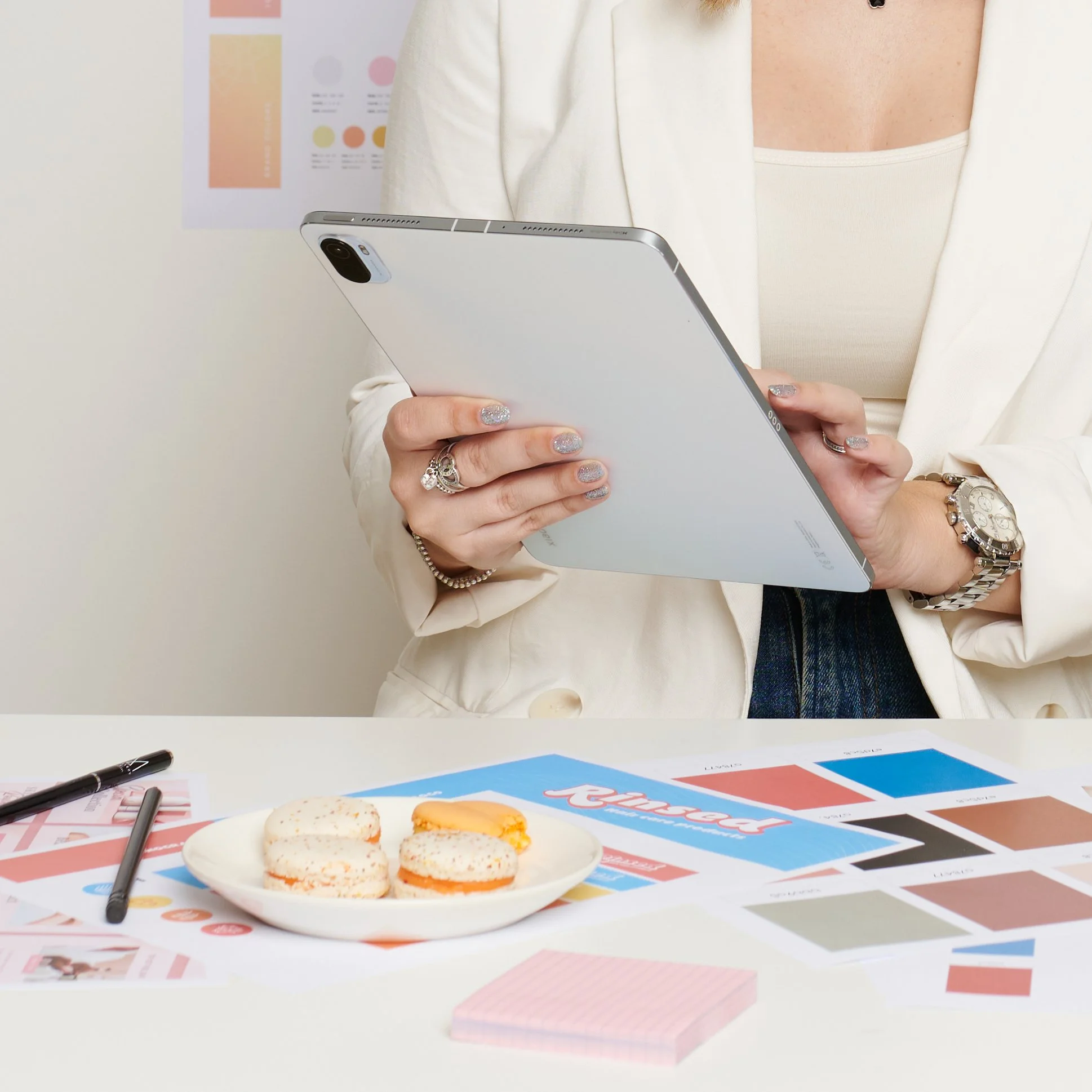 Person holding a tablet, sitting at a desk with color swatches, design prints, pens, and macarons on a plate.