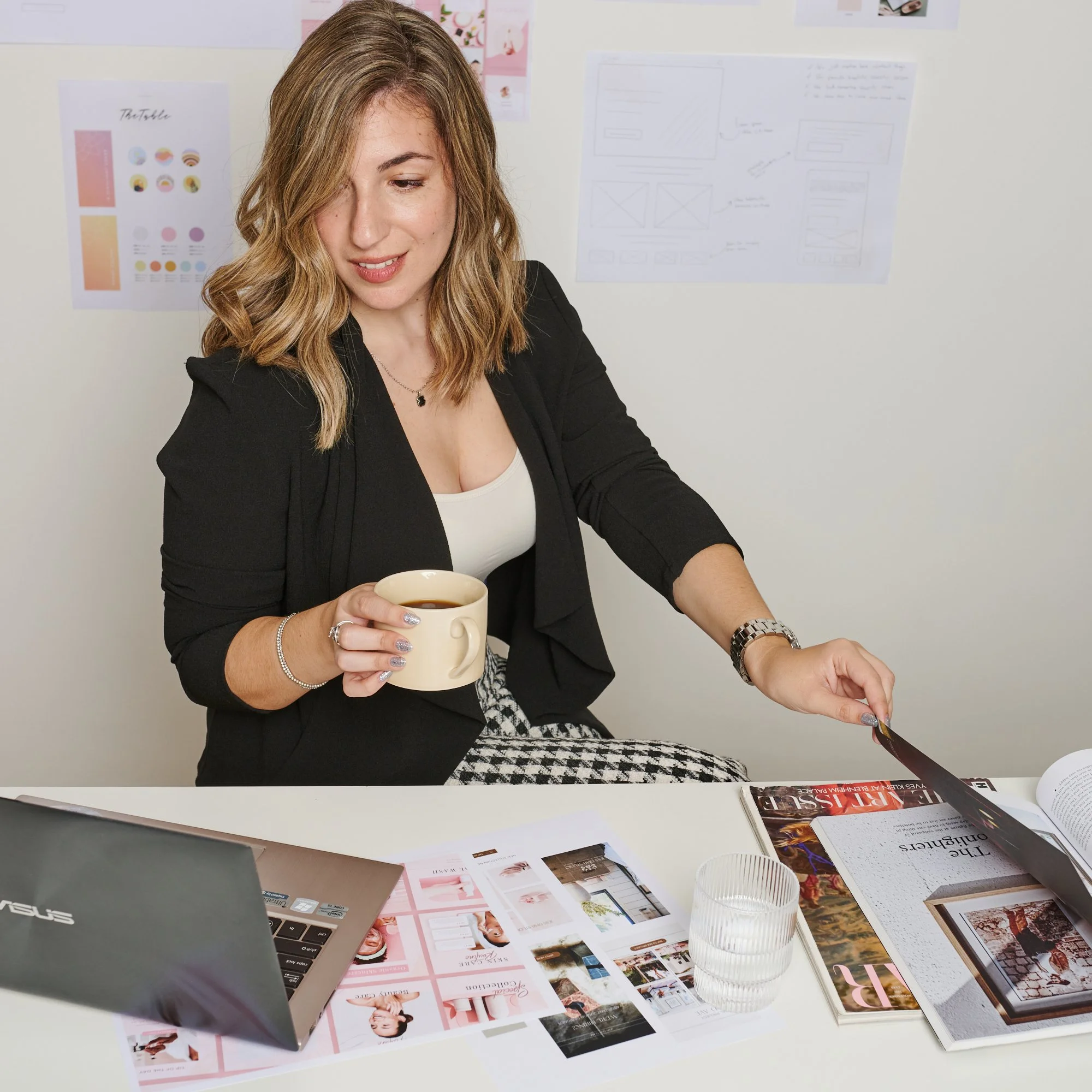 Woman in black blazer sitting at desk with laptop, holding coffee cup, and looking at magazine. Design sketches and materials on wall and table.