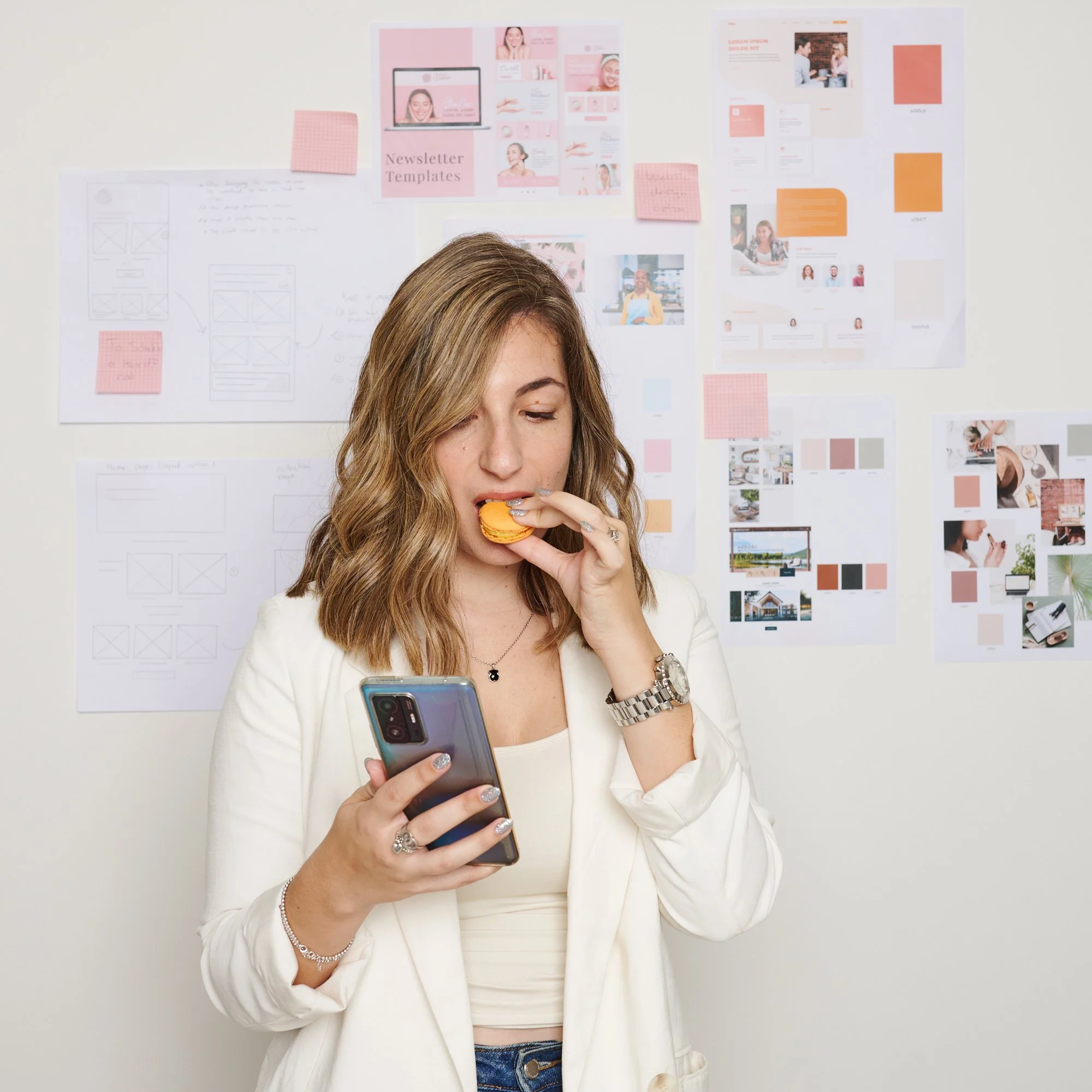 Woman in a white blazer eating a macaron and looking at her smartphone, standing in front of a wall with design templates and mood boards.