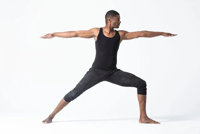 A man in athletic clothing doing a yoga warrior pose with arms extended and legs bent in a lunge stance.