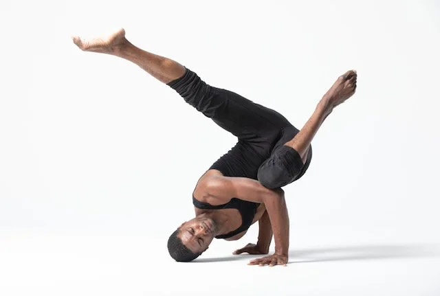 A woman performing a yoga move on a white background, balancing on one hand with her other hand and legs extended.