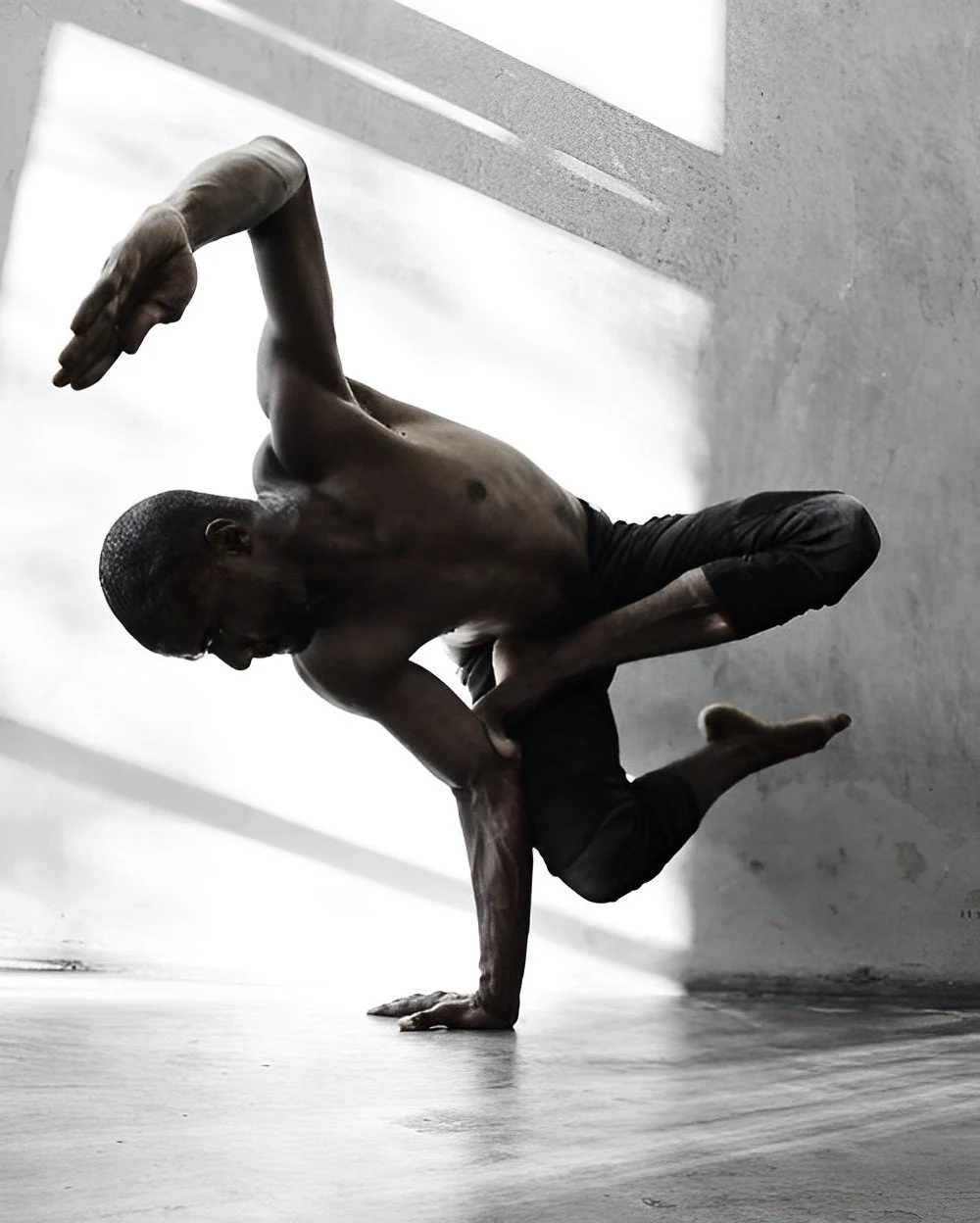 A shirtless man with glasses performing a yoga pose on one hand on a smooth floor in a minimalist room with natural light.