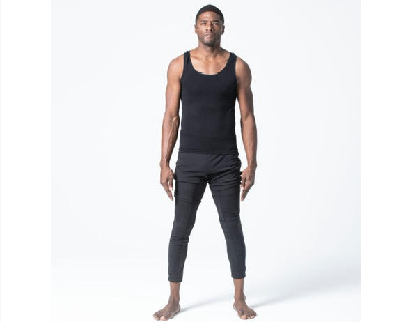 A tall man standing in a studio with a plain white background, wearing a black tank top and black pants, barefoot, looking at the camera.