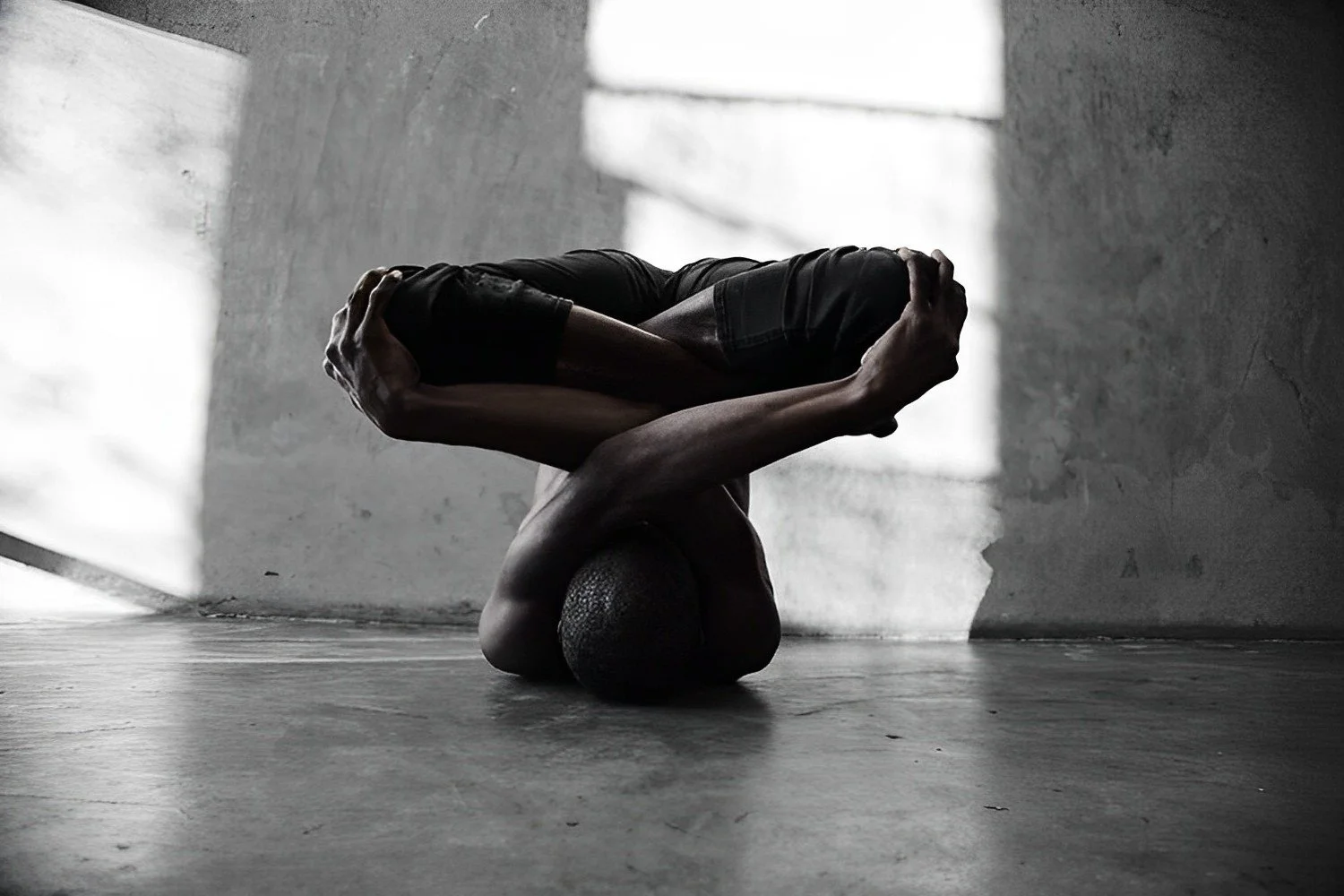A person practicing a headstand yoga pose on a textured floor in front of a concrete wall with sunlight creating shadows.