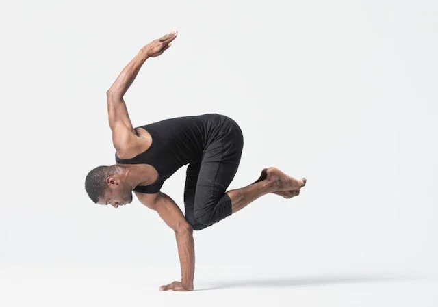 A man performing a yoga pose on a white background, balancing on one hand with his legs bent and raised.