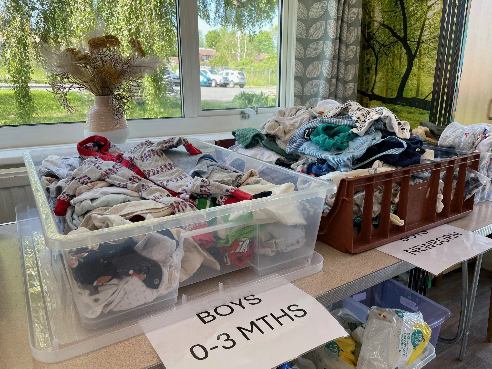 Clear plastic boxes on a table filled with sorted baby clothes labelled by age, near a window with greenery outside.