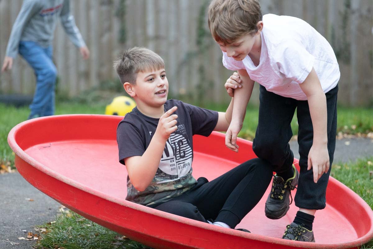 Two children playing on a red plastic tray in the garden at GL11 Community Hub