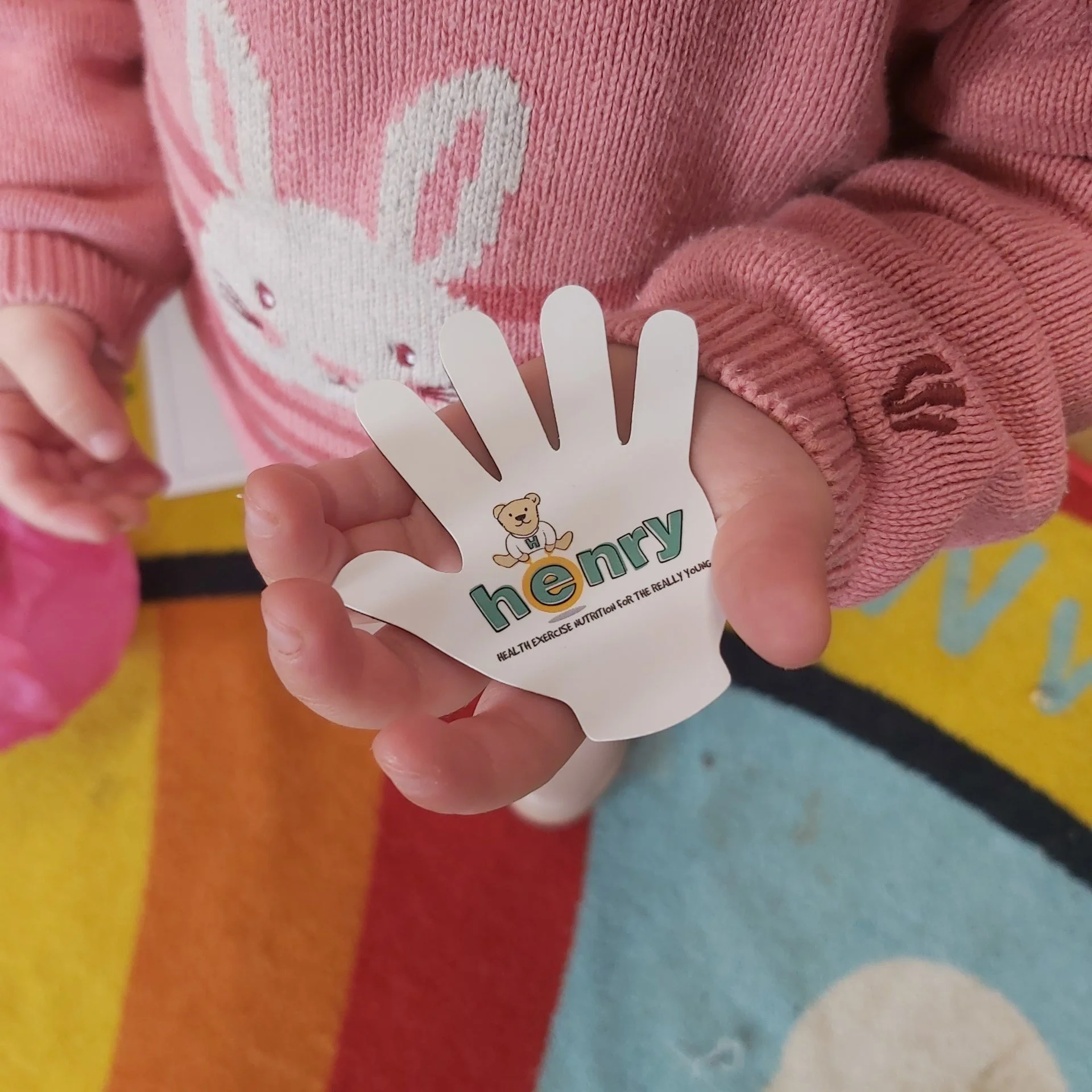 Child wearing a pink jumper with a bunny design holding a white hand-shaped sticker labelled 'henry' with a teddy bear logo.