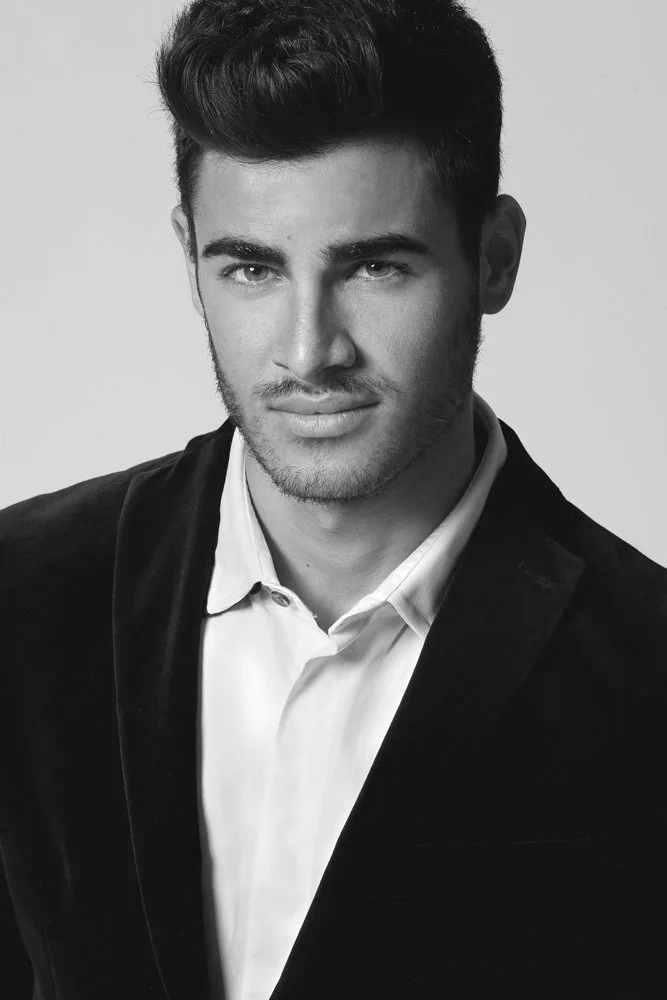 Black and white portrait of a young man with dark, styled hair, light facial hair, wearing a white shirt and a dark blazer, looking confidently at the camera.