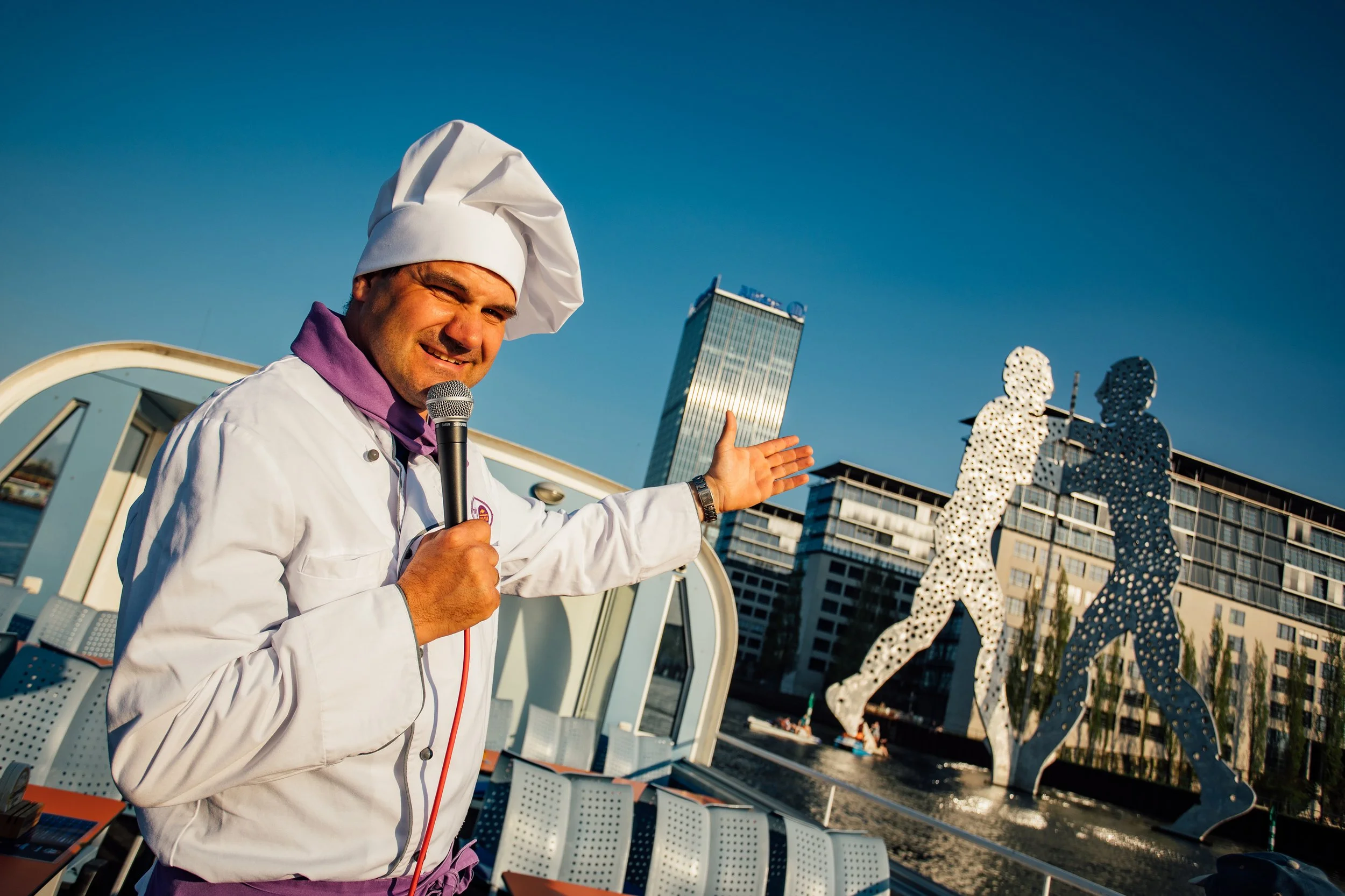 Ein Koch in weißer Uniform und Kochmütze spricht mit einem Mikrofon vor dem Scheinwerfer im Freien, mit den Skulpturen der Brüderchen im Wasser und modernen Gebäuden im Hintergrund.