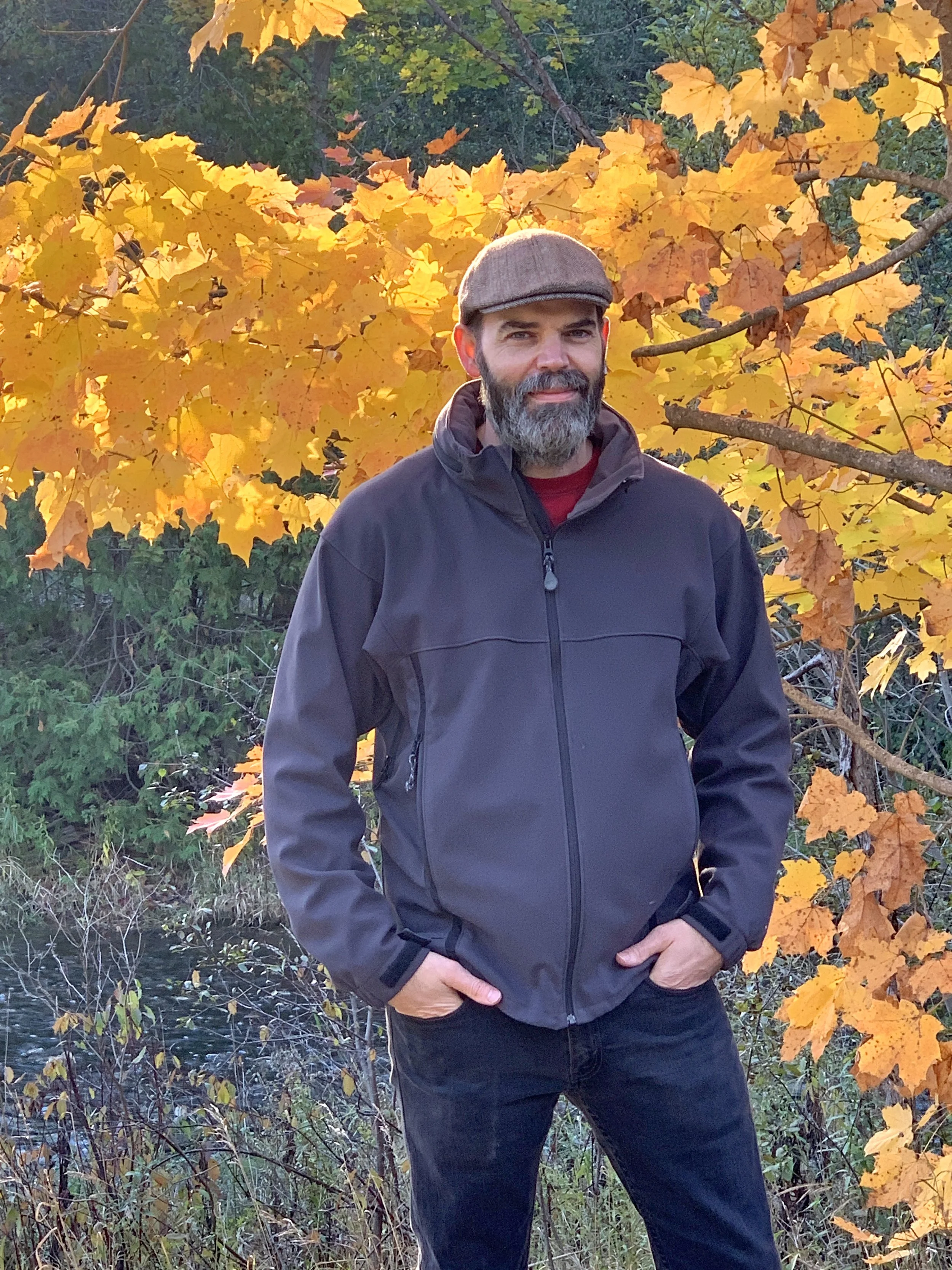 A man with a beard and mustache wearing a brown flat cap, a black jacket, and dark pants standing outdoors in front of a tree with yellow autumn leaves.