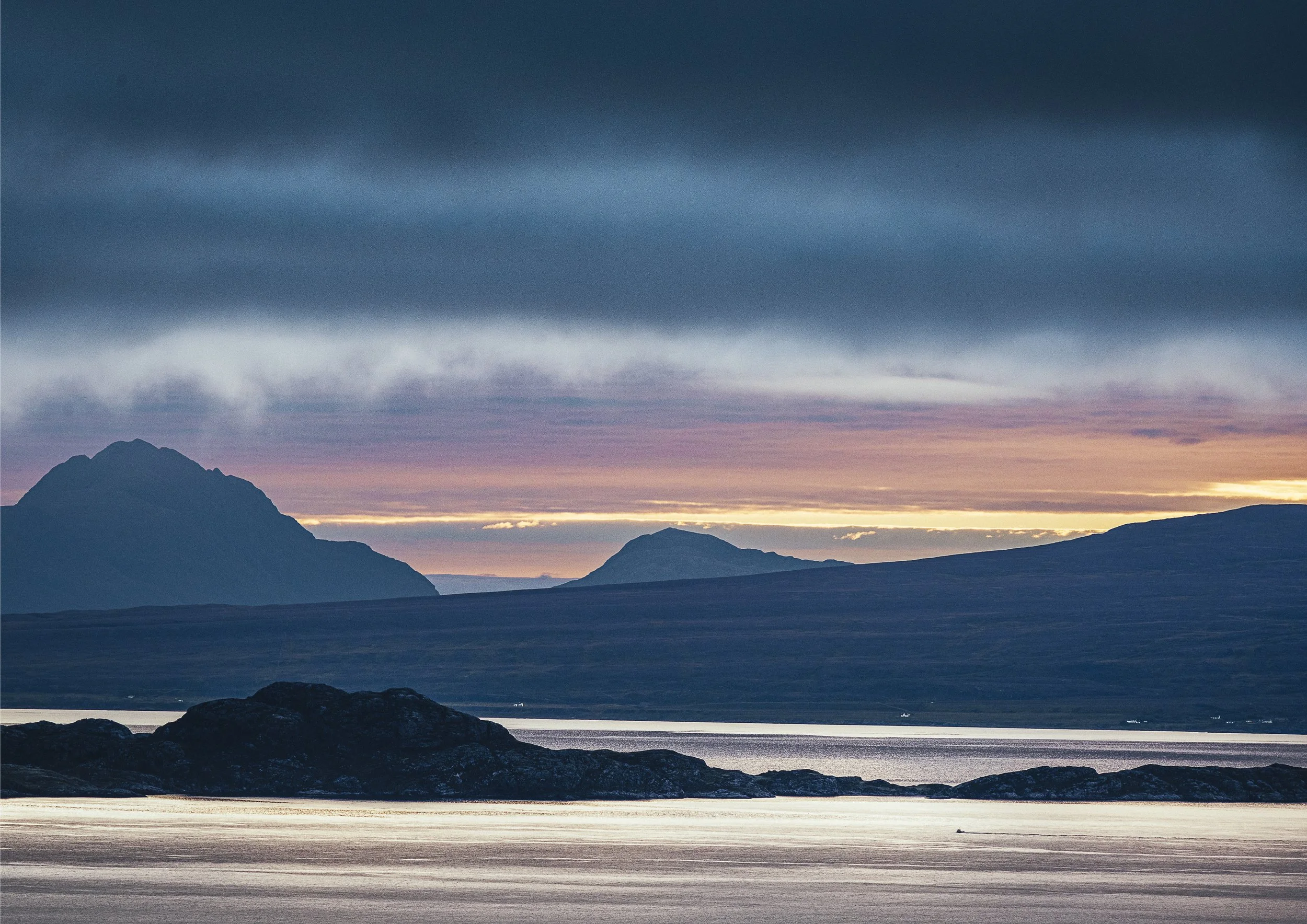 Scenic landscape of a calm lake with rocks in the foreground, rolling hills, and mountains in the background under a cloudy sky with colorful sunset hues.