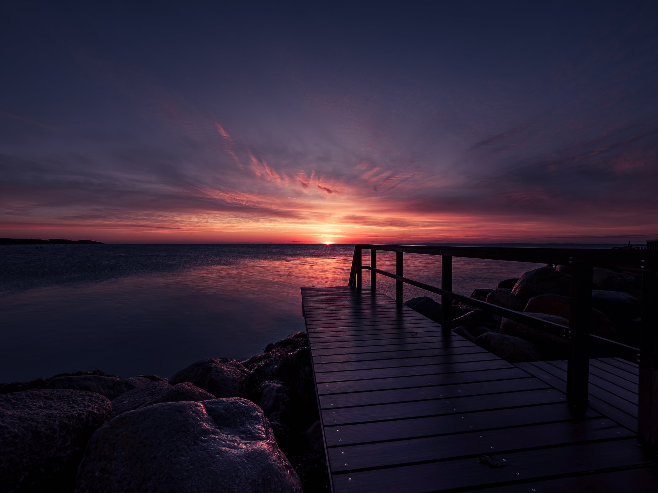 Sunset over a calm body of water with a wooden dock and rocks in the foreground, colorful sky with pink, purple, and orange clouds.