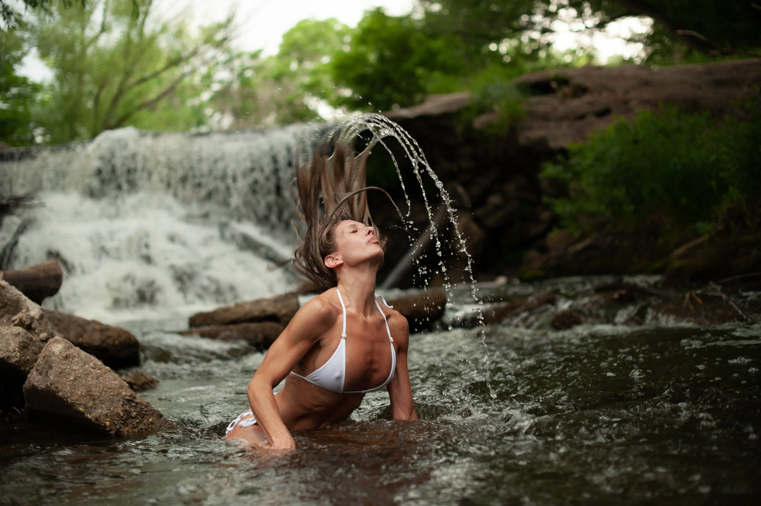 A woman in a white bikini leans back with her hair flipping in a creek near a waterfall surrounded by trees and rocks.