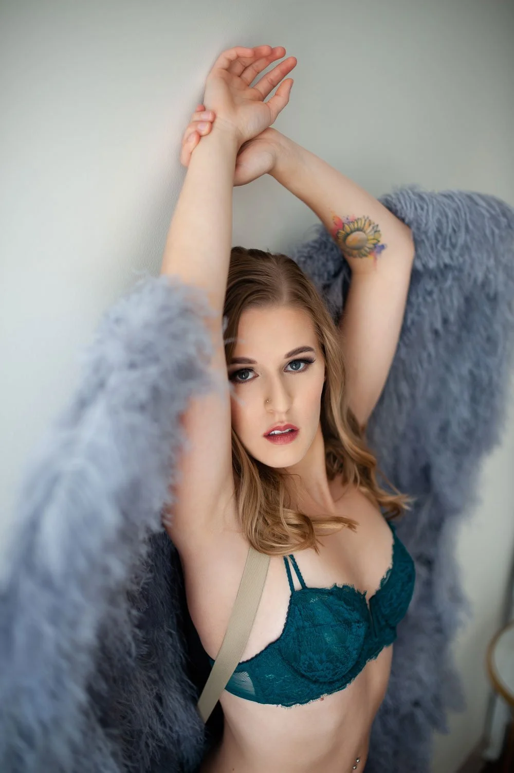 A woman with blue eyes and brown hair, wearing teal lace lingerie in grey angel wings, poses with her arms raised above her head, leaning against a wall with a tattoo on her arm.