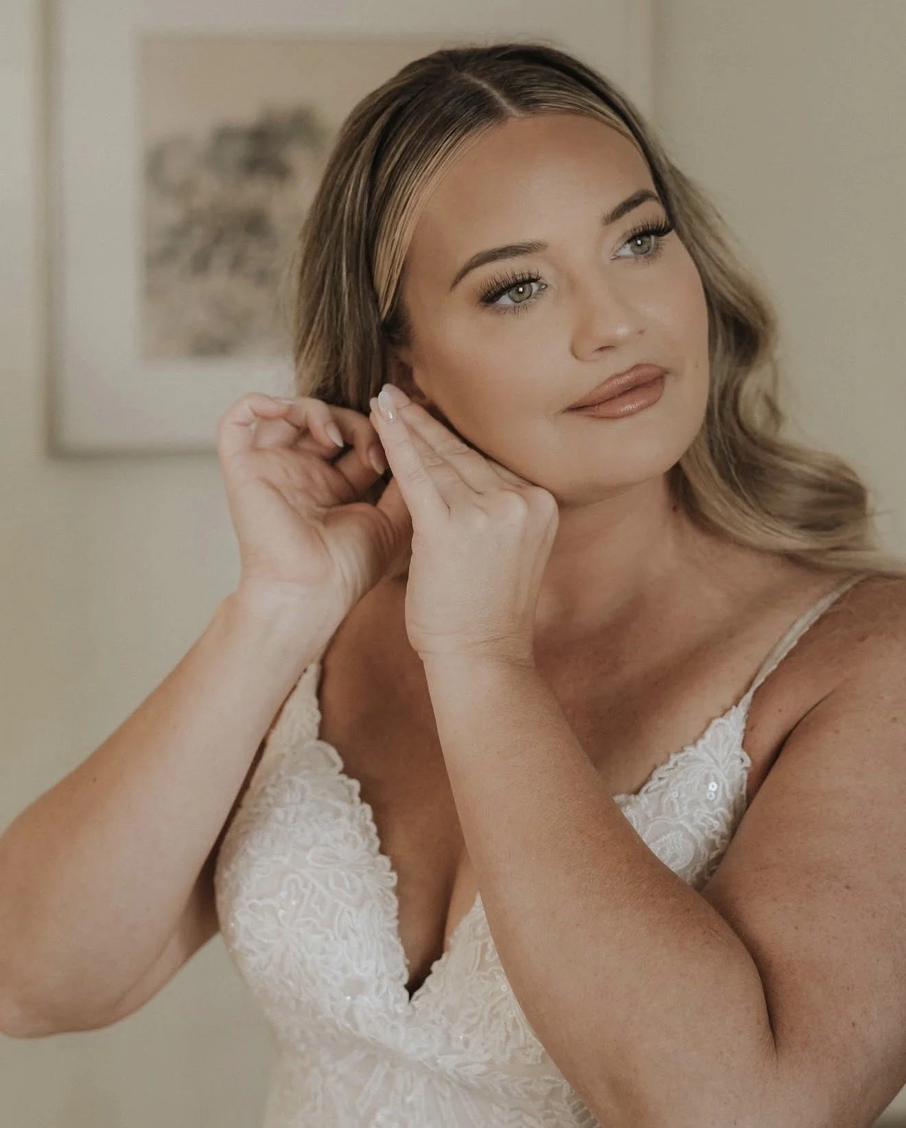 A woman in a white lace dress is touching her ear and looking at the camera with a slight smile, standing in front of a framed picture on a beige wall.