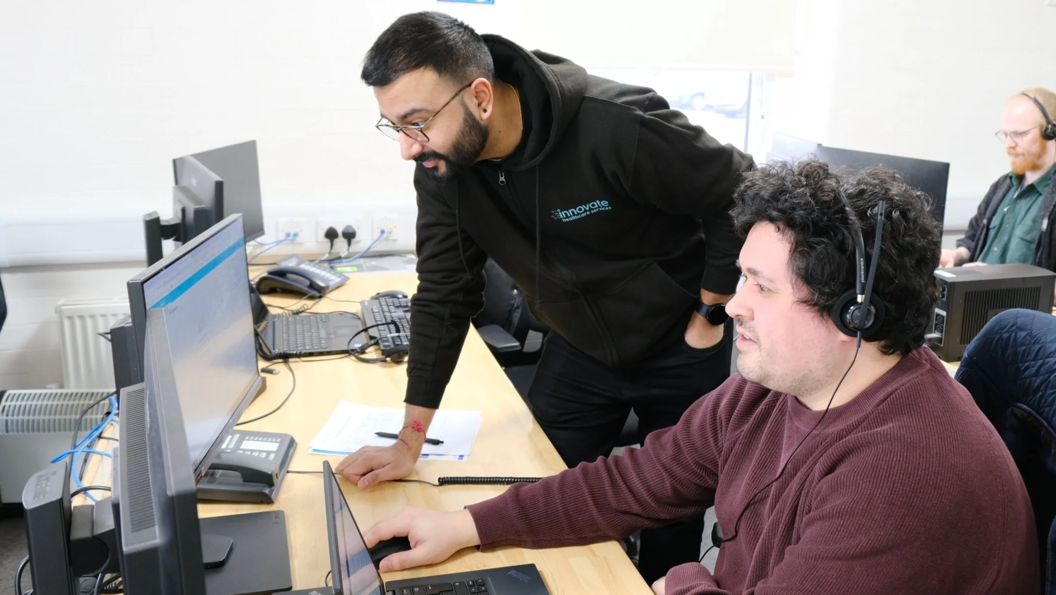 Two men working in an office, one sitting at a desk with a computer and wearing a headset, and the other standing and leaning over to look at the computer screen. The man standing is wearing a black hoodie with a logo, glasses, and has a beard. The office has multiple computers, phones, and other office equipment.