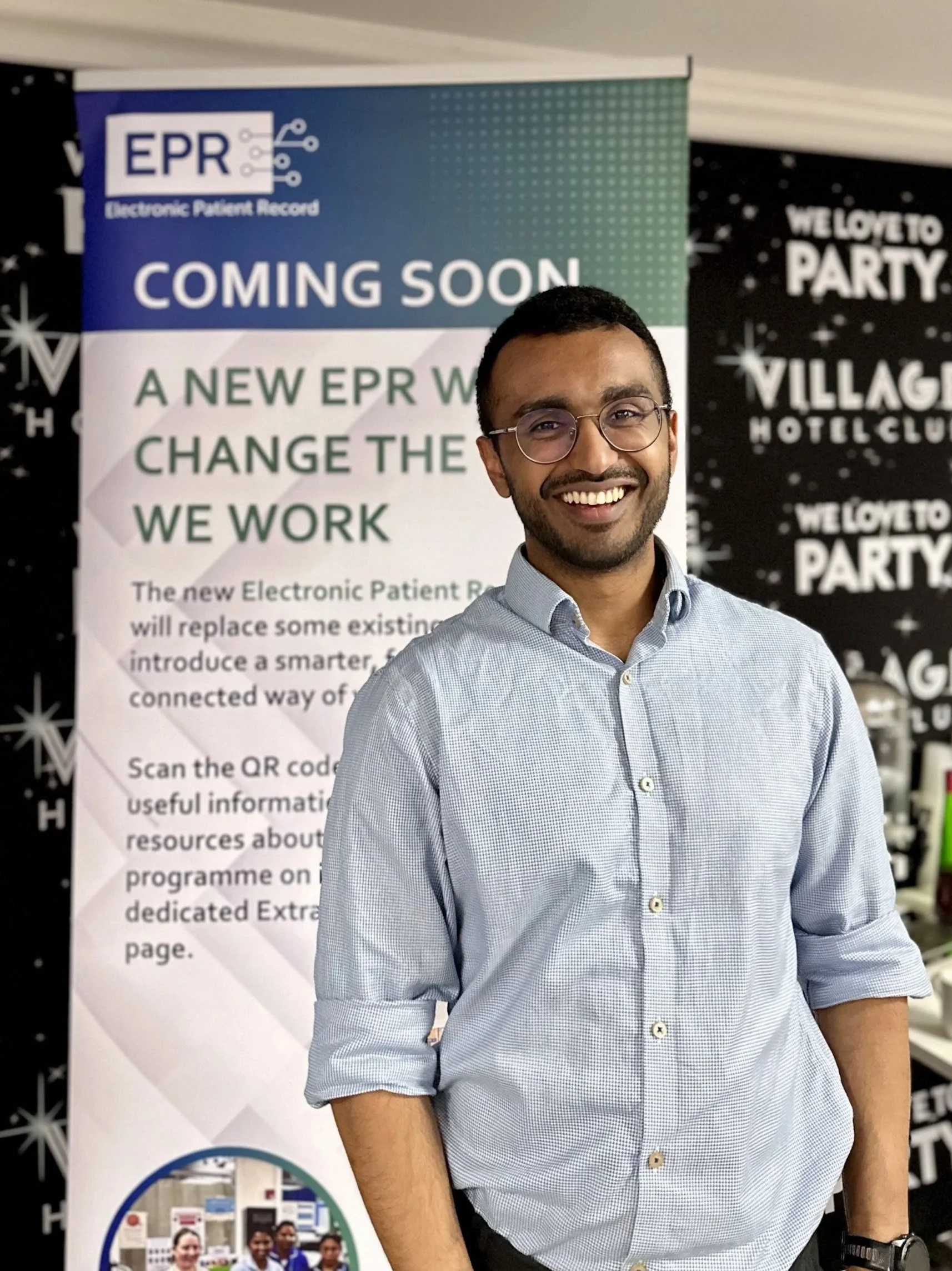 A smiling man in glasses wearing a light blue button-up shirt standing in front of a banner that reads 'Coming Soon' about a new Electronic Patient Record system.