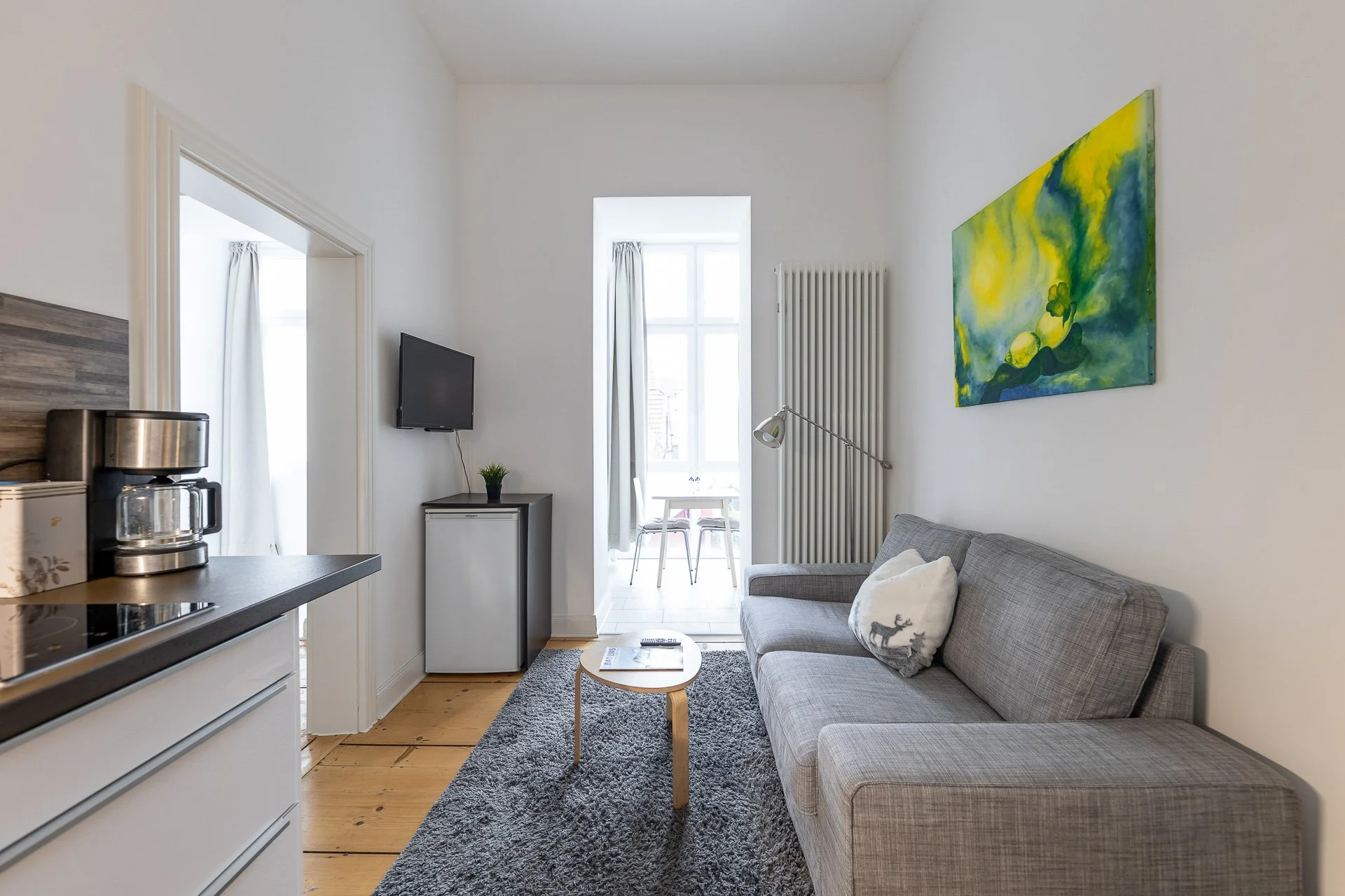 Wohnzimmer mit grauem Sofa, Couchtisch, Fernseher, Mini-Kühlschrank, Kaffeemaschine und buntem Gemälde an der Wand. Nebenraum mit Fenster im Hintergrund. Holzboden, grauer Teppich.
