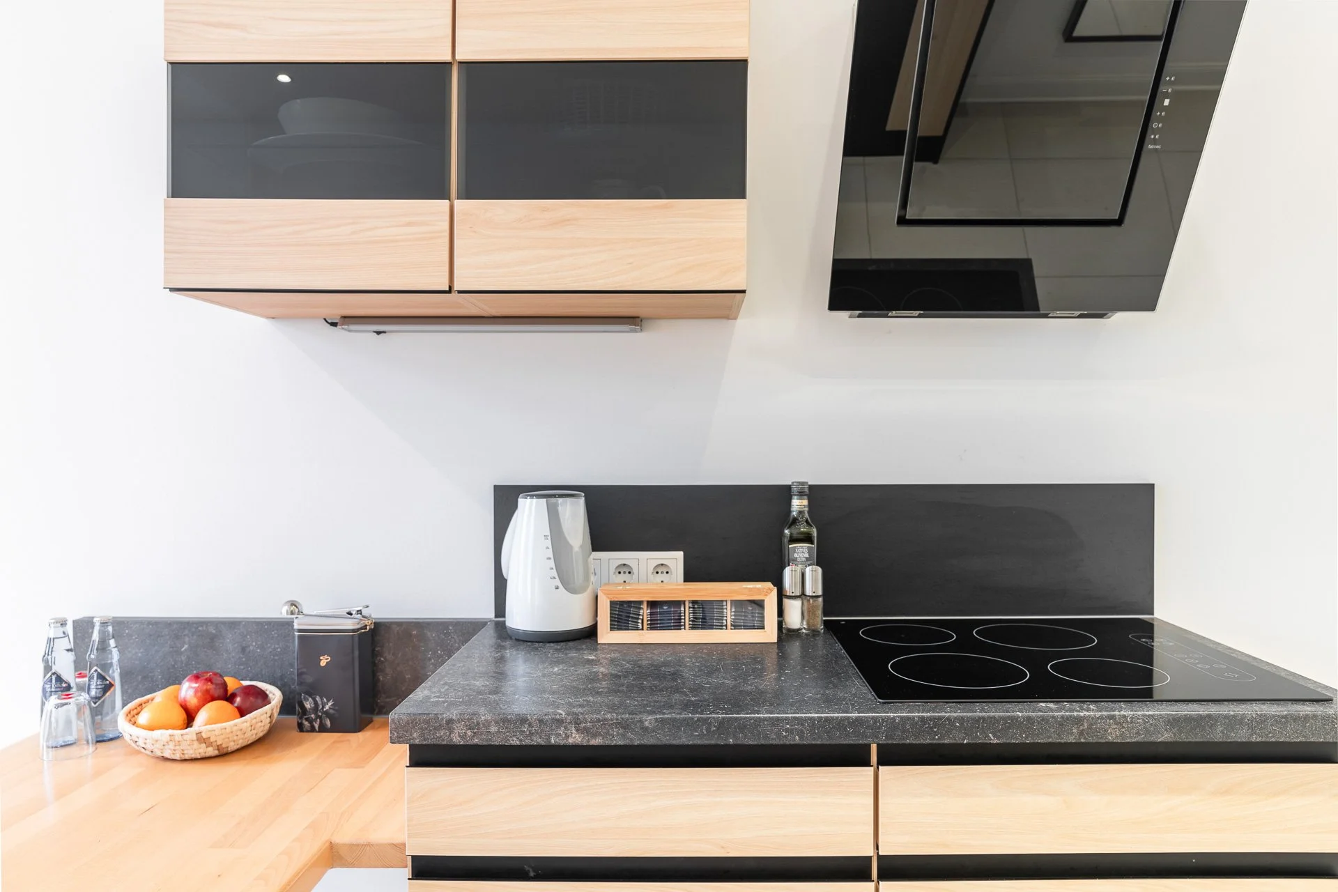 Moderne Küche mit Elektroherd, Wasserkocher, Gewürzbox, Olivenölflasche und Obstkorb auf einer hölzernen Arbeitsplatte.