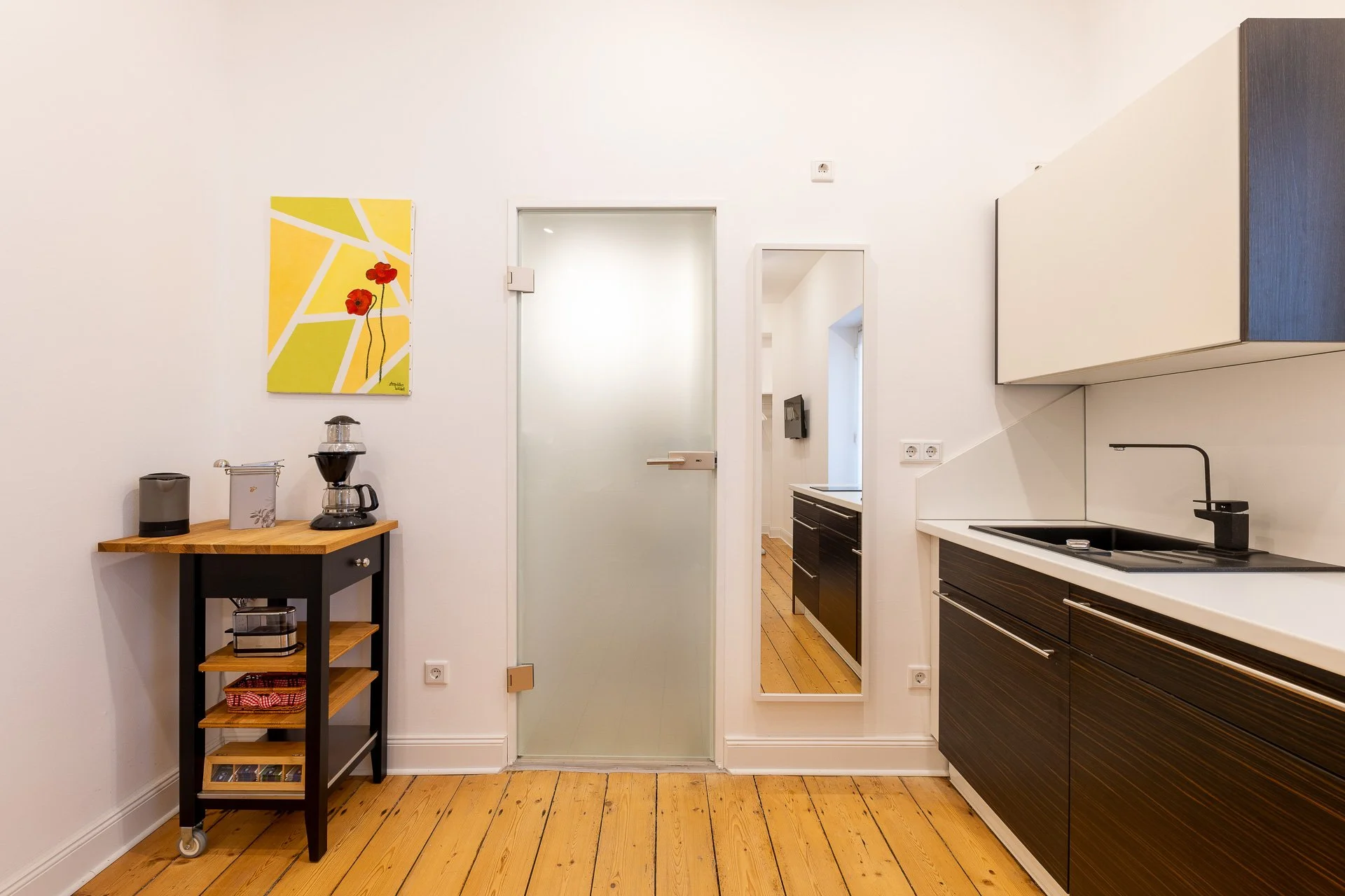 Moderne Küche mit Holzboden, Küchentheke mit Wasserkocher und Kaffeemaschine, Glas-/Glastür, Spiegel an der Wand, Bild mit roten Blumen, schwarze und weiße Schränke, schwarzes Spülbecken.