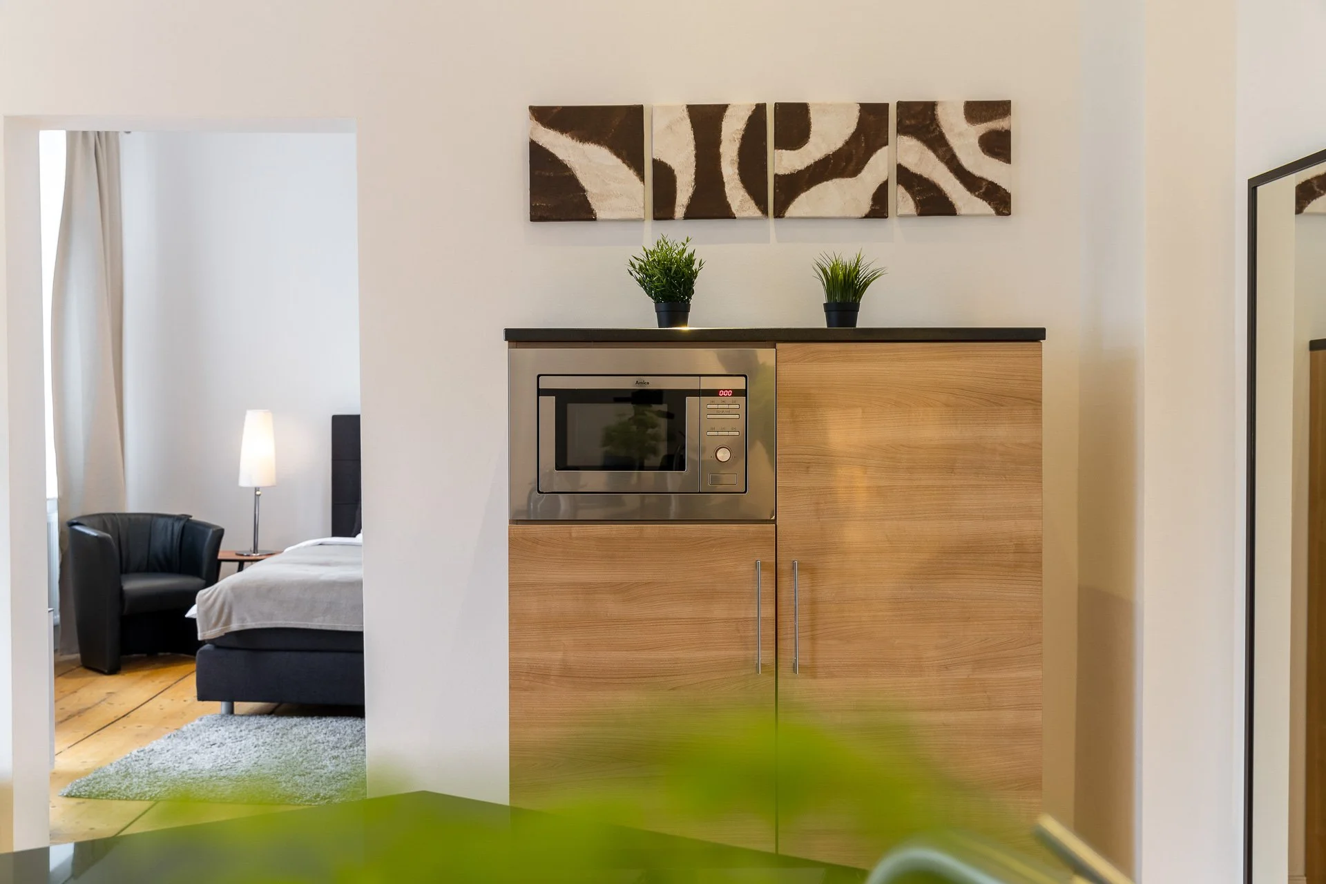 Innenraum mit Holzmöbeln, Mikrowelle, zwei kleinen Pflanzen, abstrakter Kunst an der Wand und Blick ins Schlafzimmer mit Bett, Sessel und Lampe.