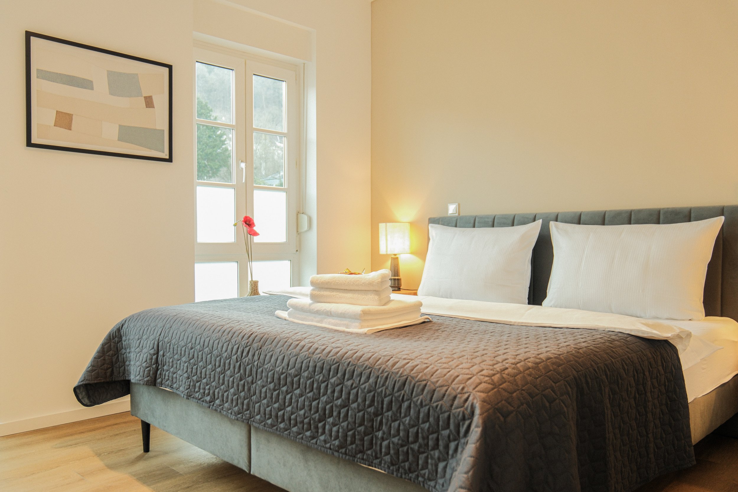 Modernes Schlafzimmer mit Doppelbett, grauer Tagesdecke, gestapelten weißen Handtüchern, Nachttischlampe, Wandkunst und Fenster.