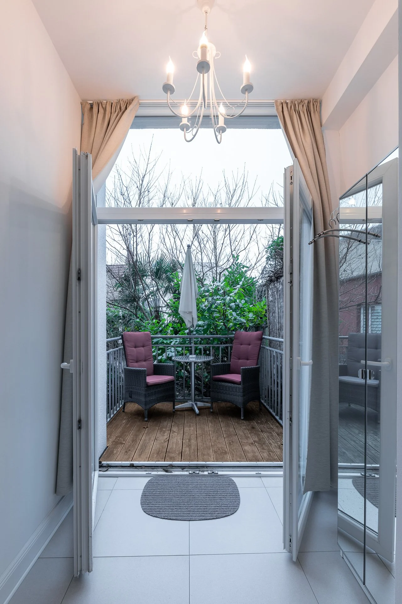 Blick vom Innenraum auf einen Balkon mit offenem Doppelflügeltüren, zwei Rattansessel mit roten Polstern, einem kleinen Tisch und einem zusammengefalteten Sonnenschirm. Eine Hängelampe hängt im Innenraum, Vorhänge sind aufgezogen und im Hintergrund s