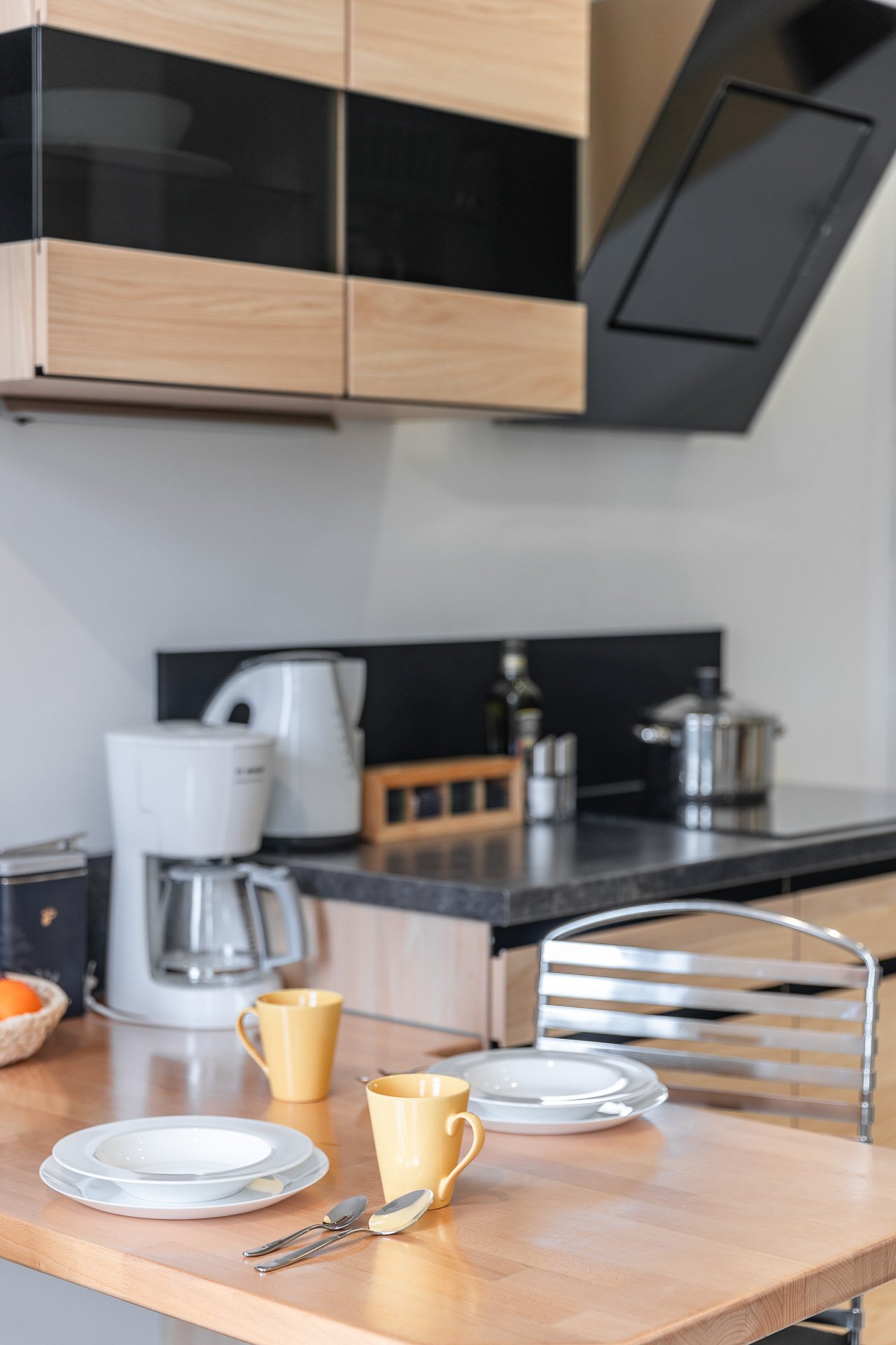 Moderne Küche mit Teller, Tassen, Besteck auf einem Holztisch, Küchenzeile mit Kaffeemaschine, Gewürzregal und Topf.