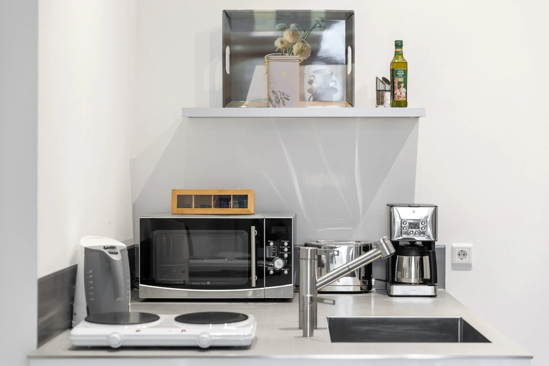 Moderne Küchenecke mit Mikrowelle, Kaffeemaschine, Wasserkocher, Ölflasche und Spüle.