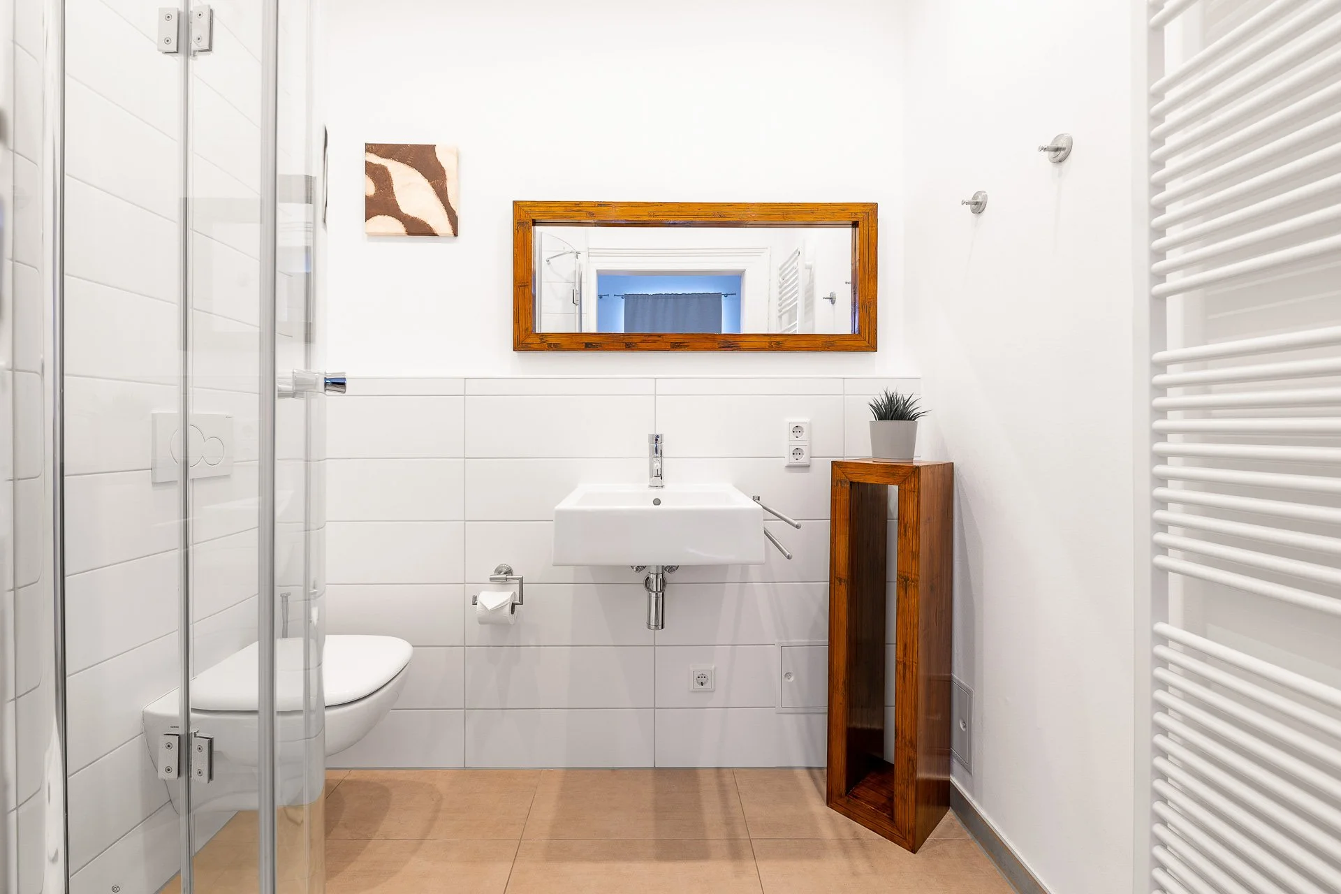 Modernes Badezimmer mit weißer Keramiktoilette, Waschbecken, rechteckigem Spiegel mit Holzrahmen, Duschkabine, beige Fliesenboden, Handtuchhalter und kleinem Pflanzenarrangement.