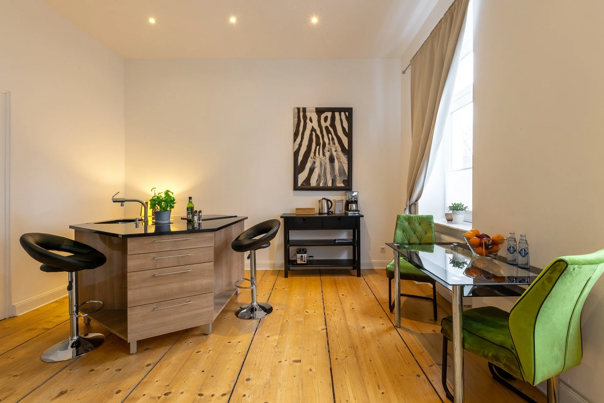 Moderne Küche mit Holzboden, Kücheninsel mit schwarzer Arbeitsplatte, zwei Barhockern, Tisch mit grünen Stühlen, Zimmerpflanze, Obstschale und Kaffeemaschine. Ein Zebra-Gemälde hängt an der Wand.