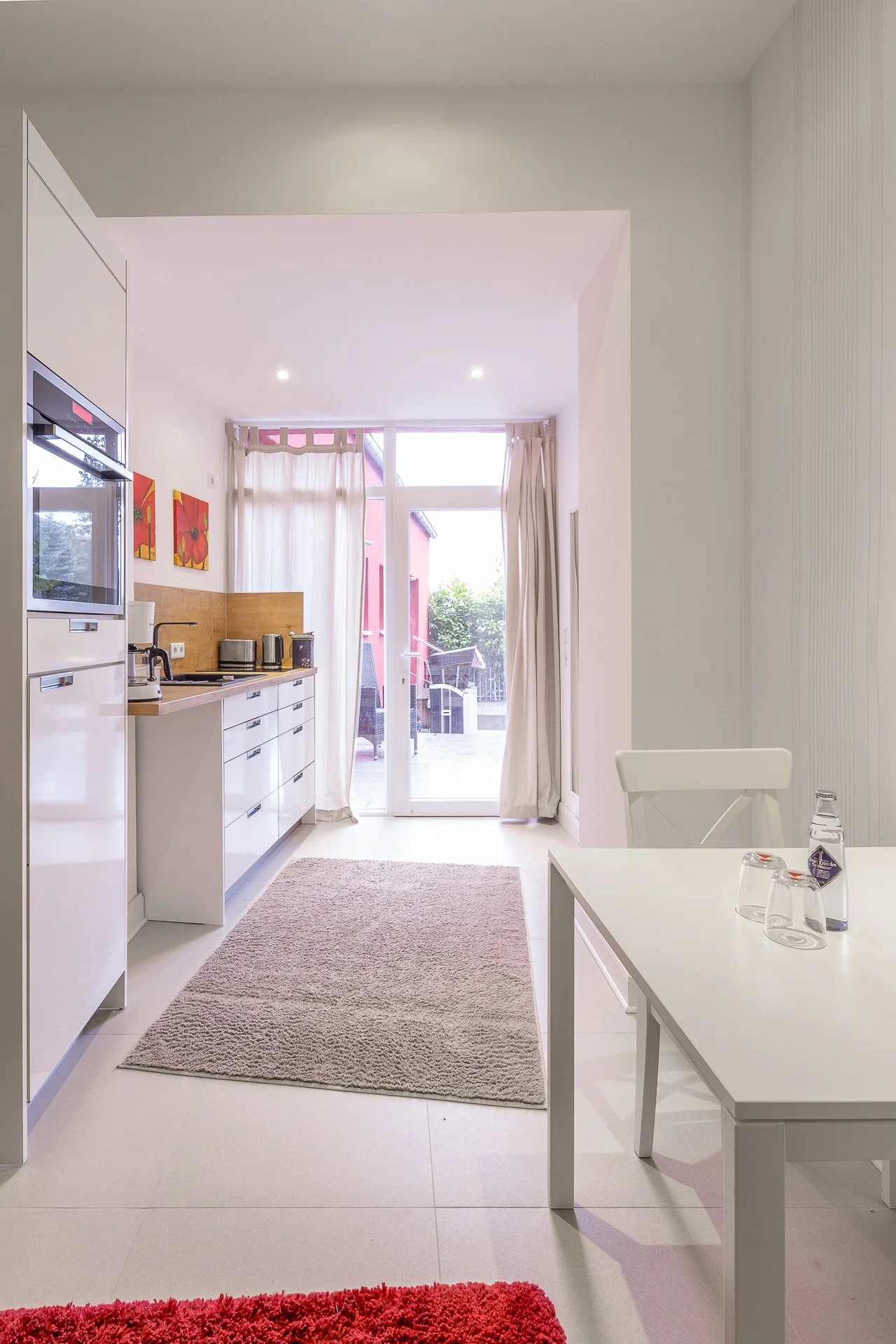 Moderne Küche mit weißem Mobiliar, Tisch mit Glasflaschen und Gläsern, elektrische Geräte, Teppich, großer Fensterbereich mit Gardinen, Zugang zur Terrasse im Hintergrund.