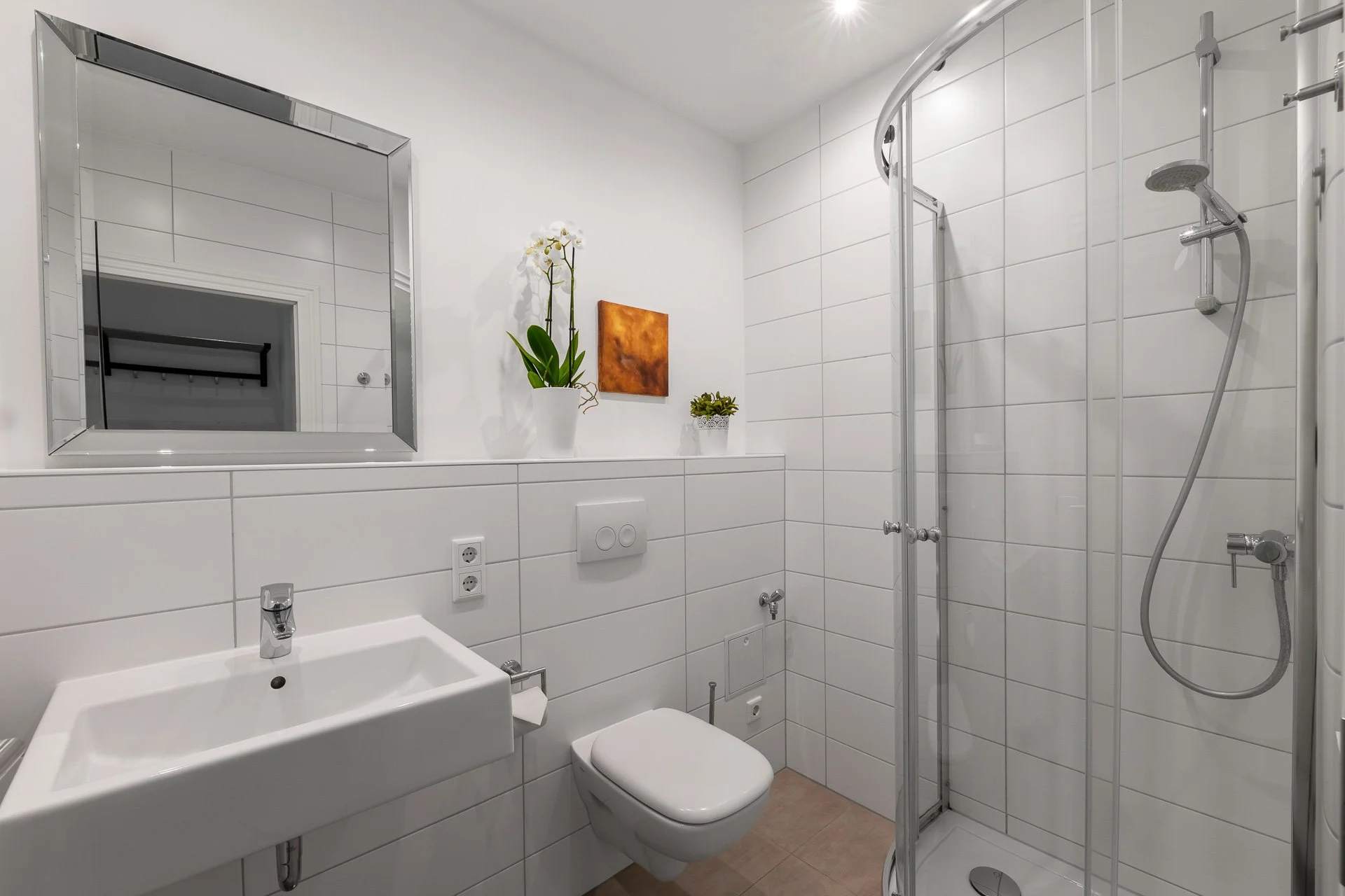 Modernes Badezimmer mit weiß gefliesten Wänden, quadratischem Spiegel über einem Waschbecken, Hänge-WC, Glasdusche, Orchidee in einem Topf und kleinem Gemälde an der Wand.