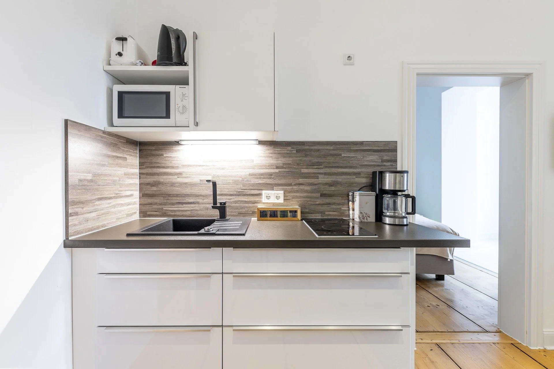 Moderne Küchenzeile mit weißem Schrank, Mikrowelle, Wasserkocher, Spüle, Kaffeemaschine und Herd. Holzfußboden im Hintergrund.